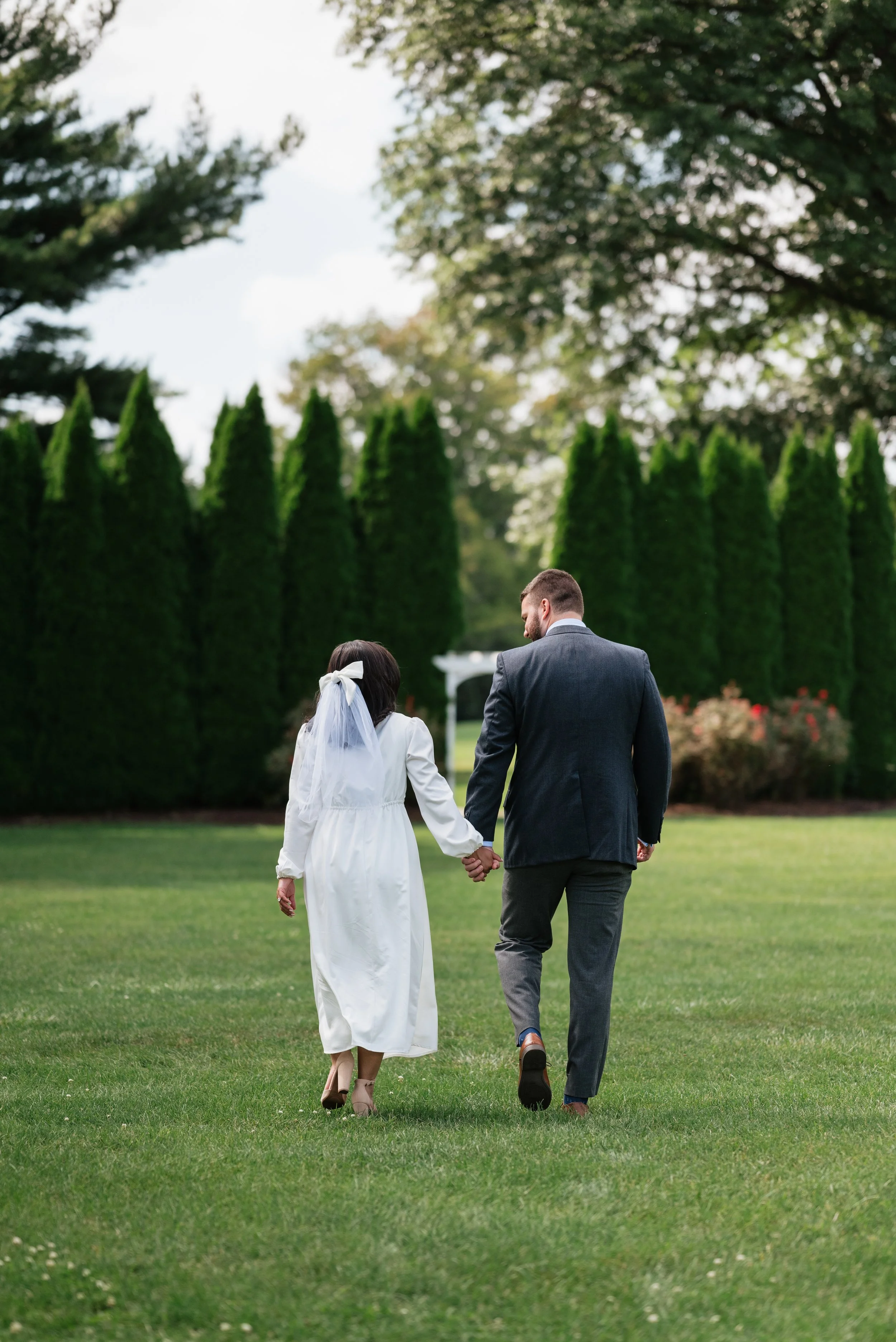 A couple, a woman in a white dress and veil and a man in a suit, walking hand in hand on a green lawn with trees in the background.