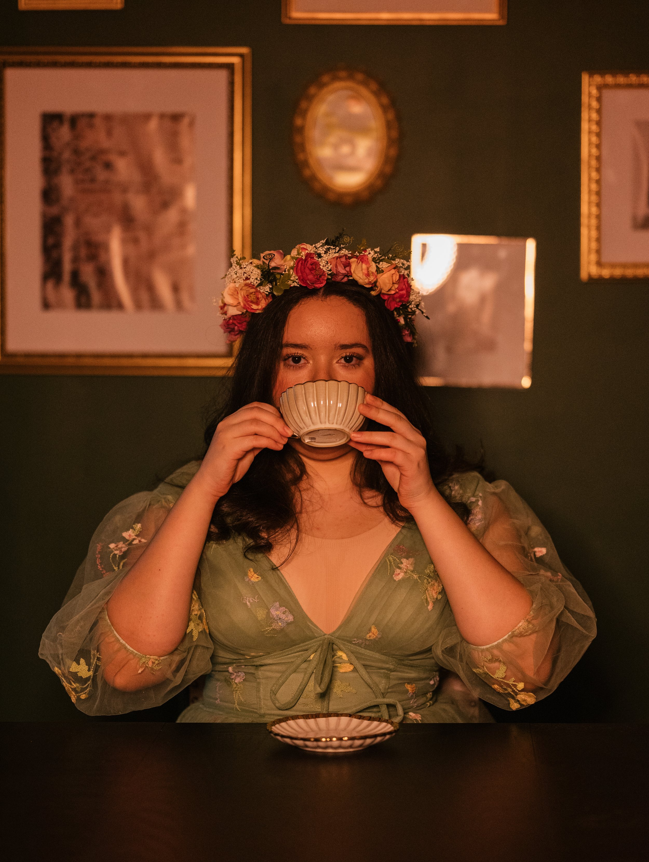 A woman with dark hair wearing a floral crown and a sheer, embroidered green top is sitting at a table in a dimly lit room. She is holding a teacup up to her face and looking directly at the camera, with a pink and gold bowl placed on the table in fr