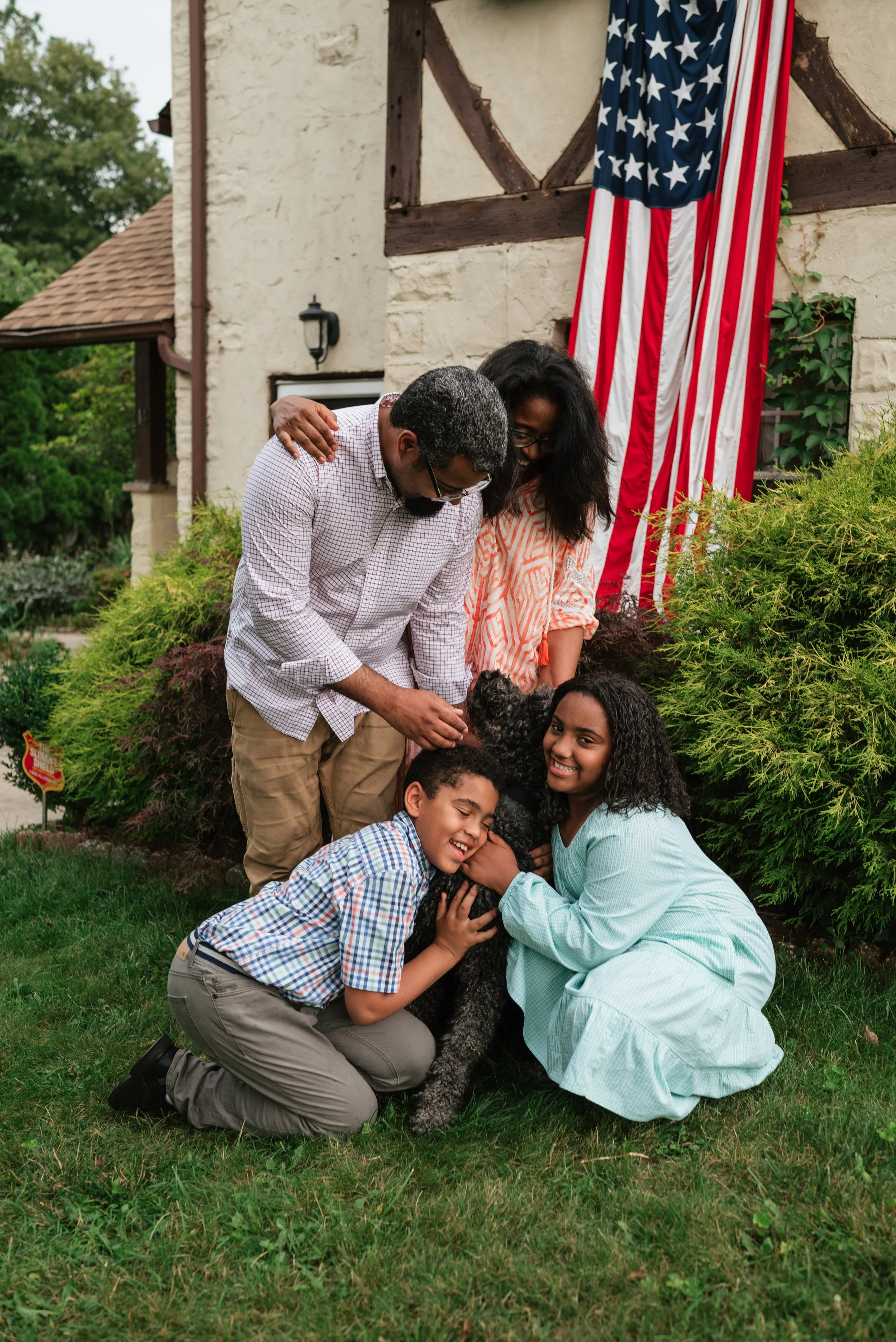 A family of five, including two smiling children and a large poodle, stands together in front of a house with an American flag. They are all interacting happily with the dog and each other, surrounded by lush greenery.