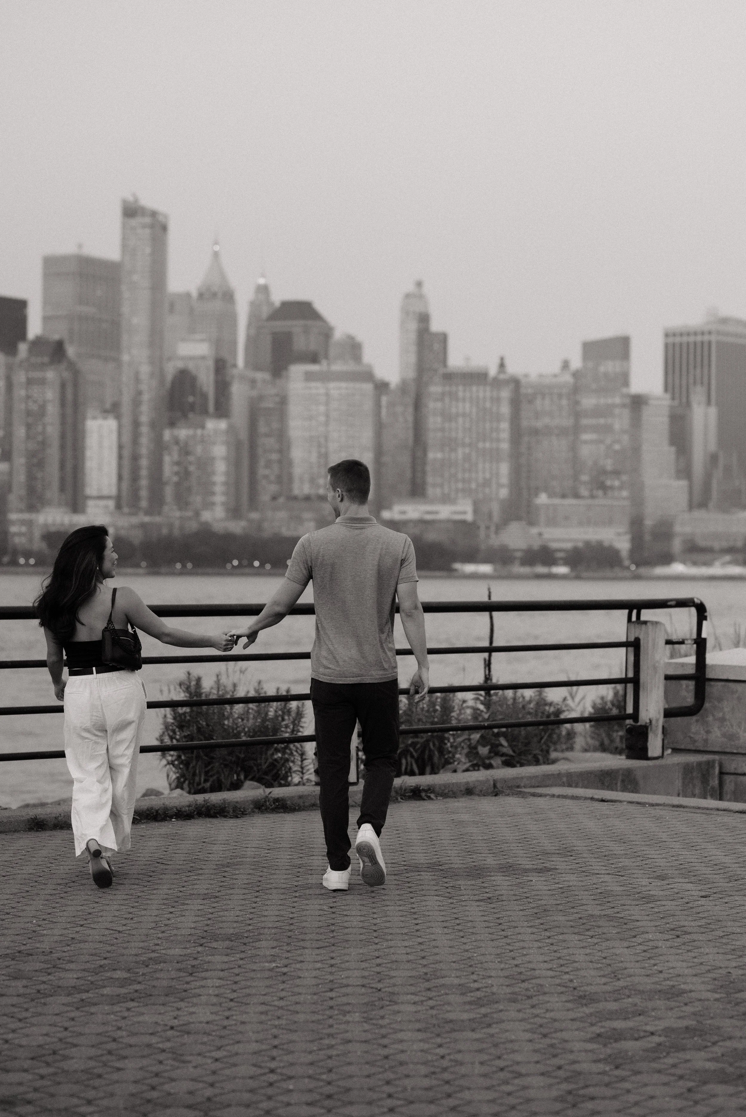 A black and white photo of a man and woman holding hands as they walk along a waterfront with a city skyline in the background.
