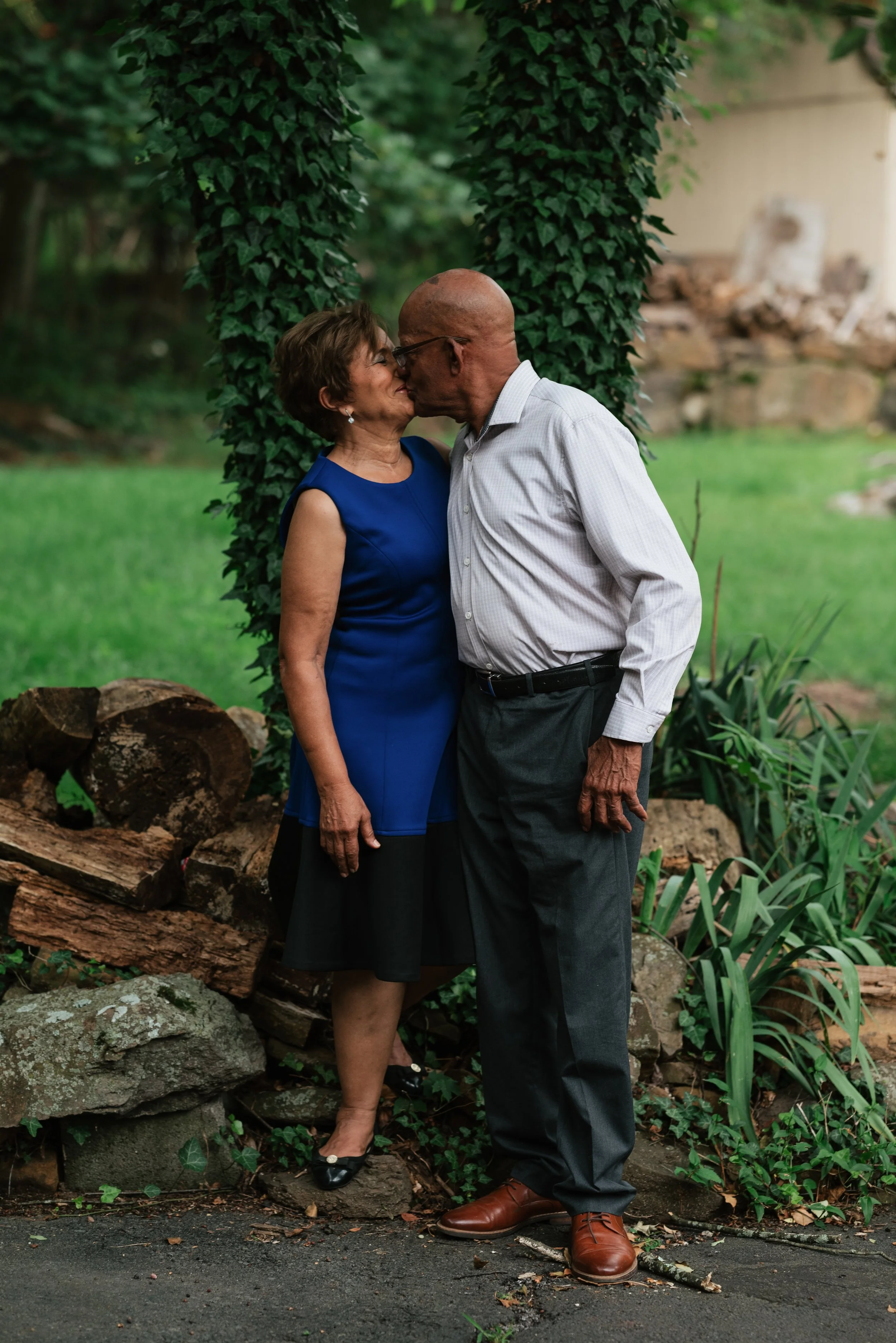 An elderly couple standing close and kissing outdoors in a garden near a tree with leafy vines.
