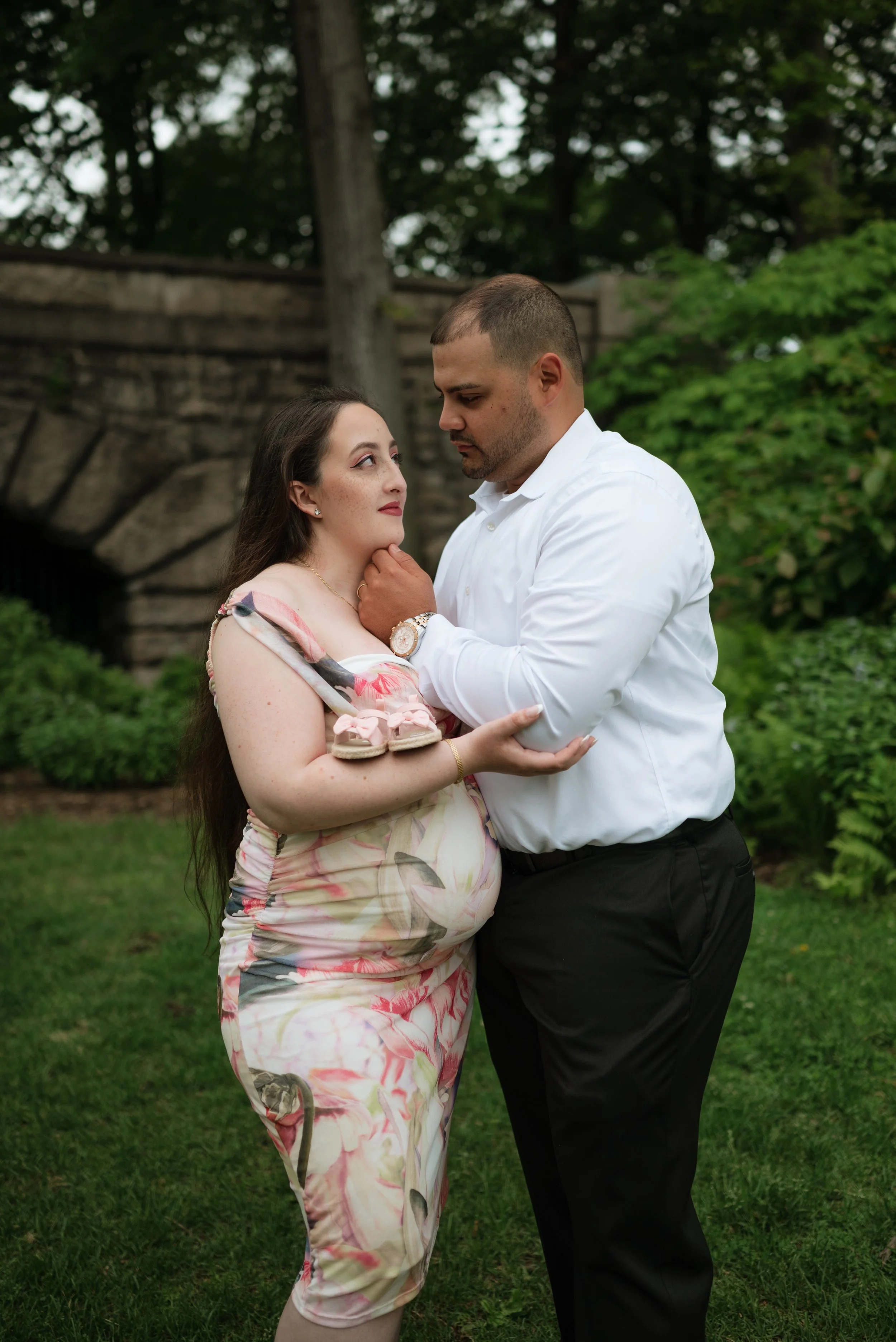 A man and a woman are standing close together outdoors, with the woman holding a pair of baby shoes. The man gently touches the woman's chin, and they look into each other's eyes. There is greenery and a stone bridge in the background.
