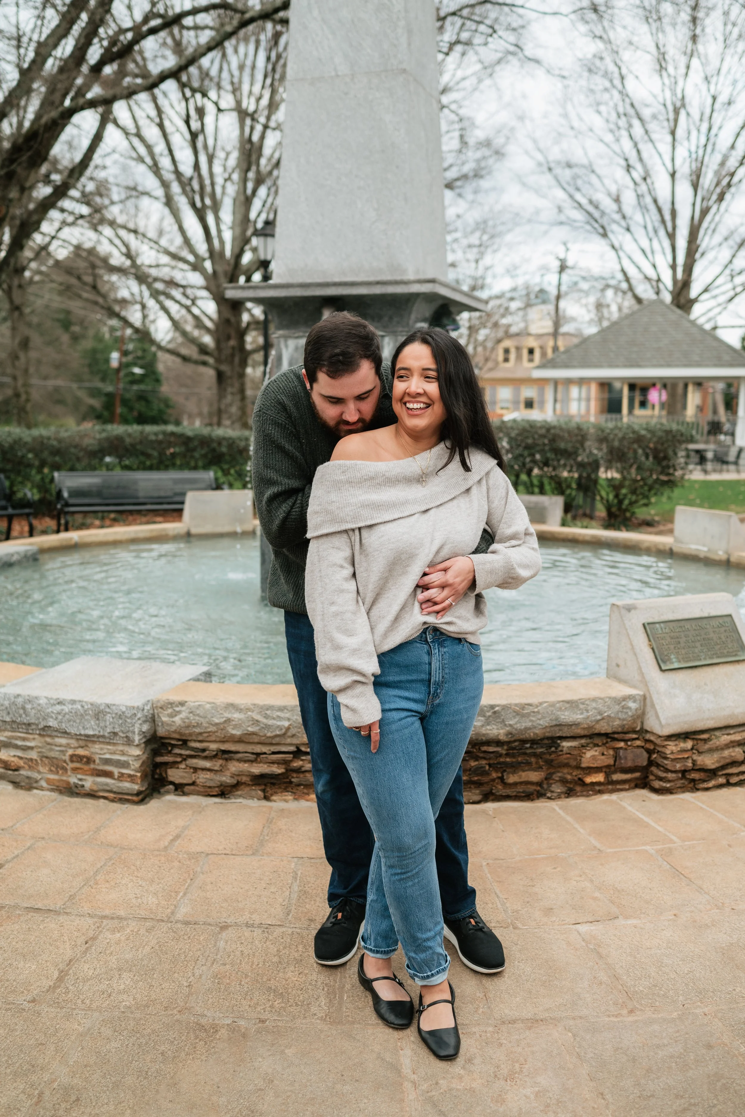 A couple standing in front of a statue and fountain in a park, laughing and embracing, with trees and houses in the background.