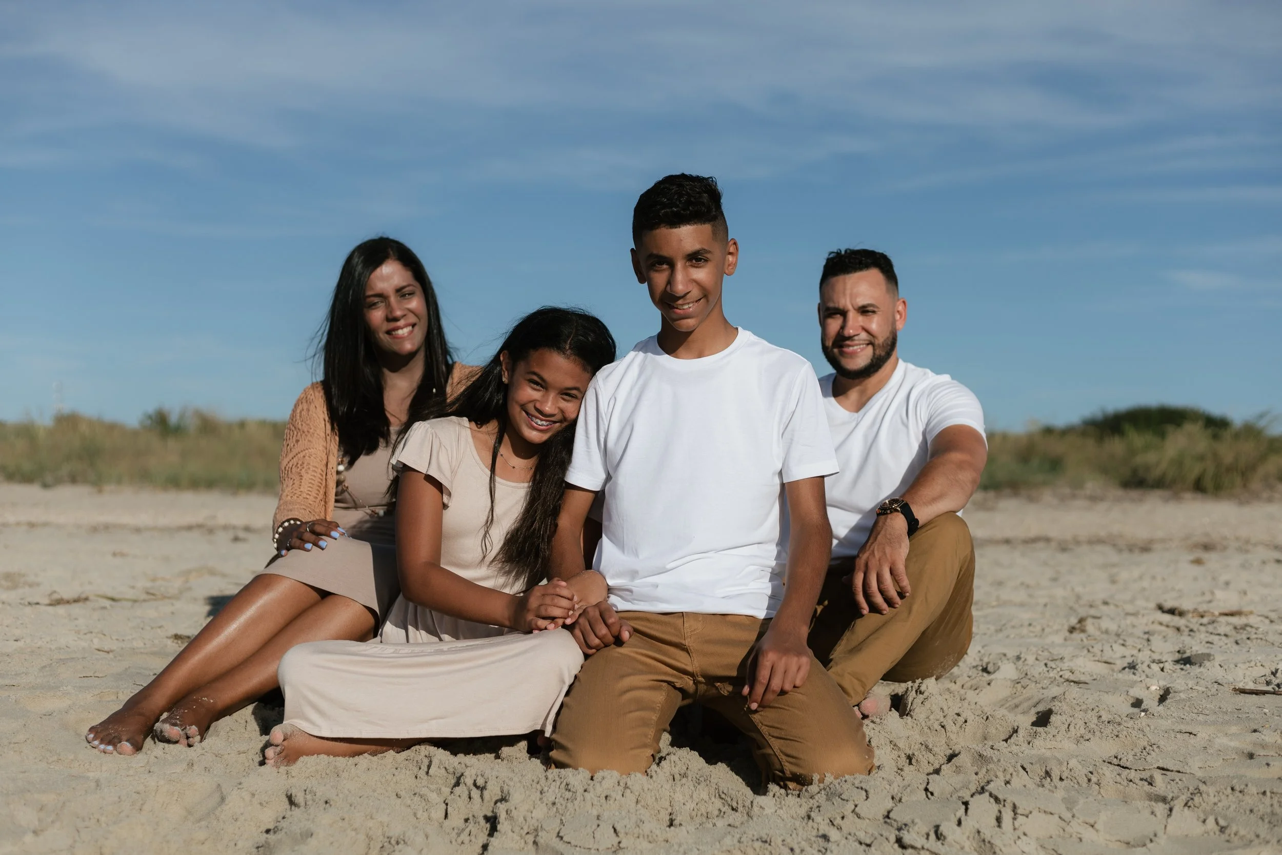 Group of four smiling people sitting on the sandy beach under a blue sky.