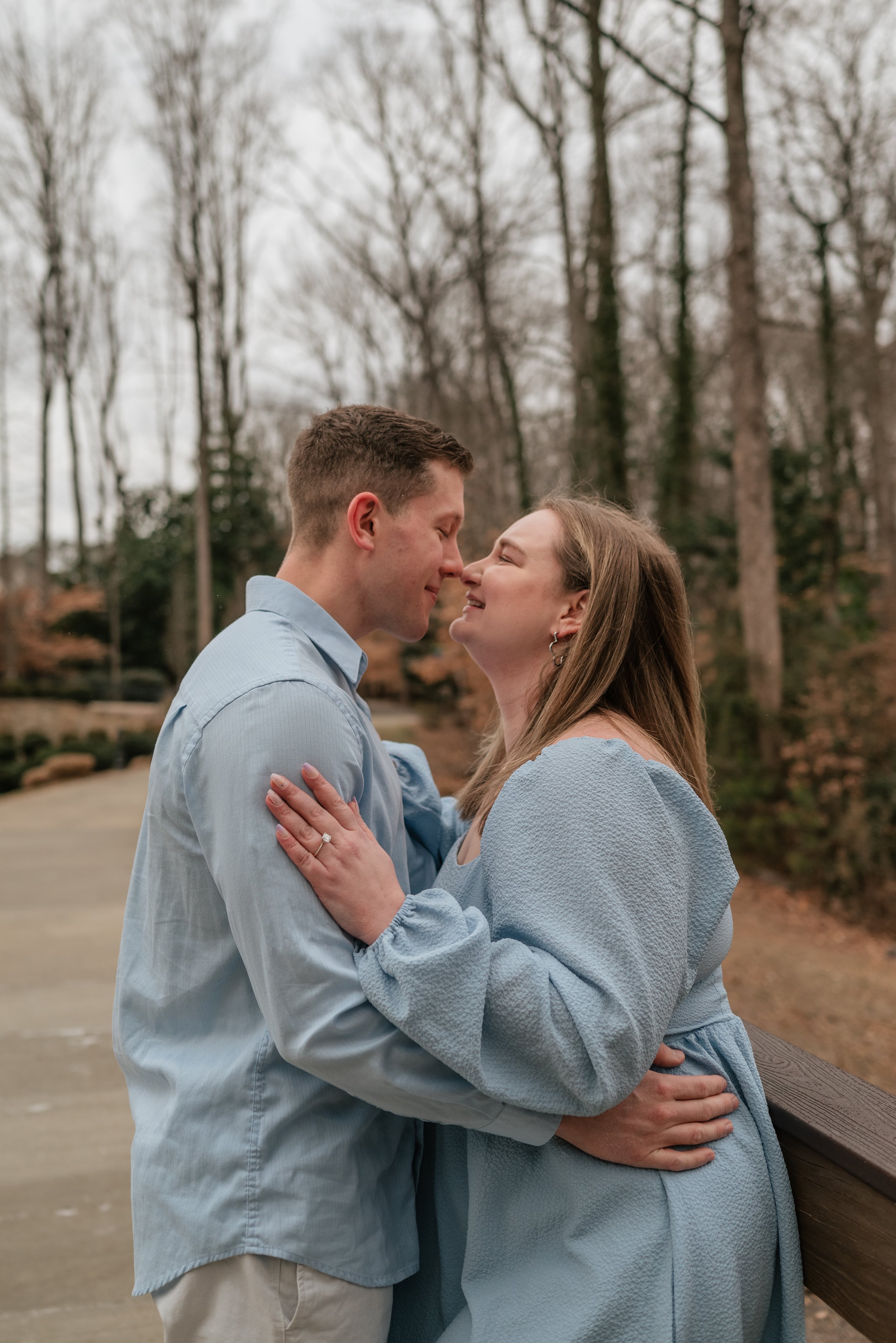 A couple standing close together outdoors, with noses touching, embracing each other and smiling, in a wooded park during autumn.