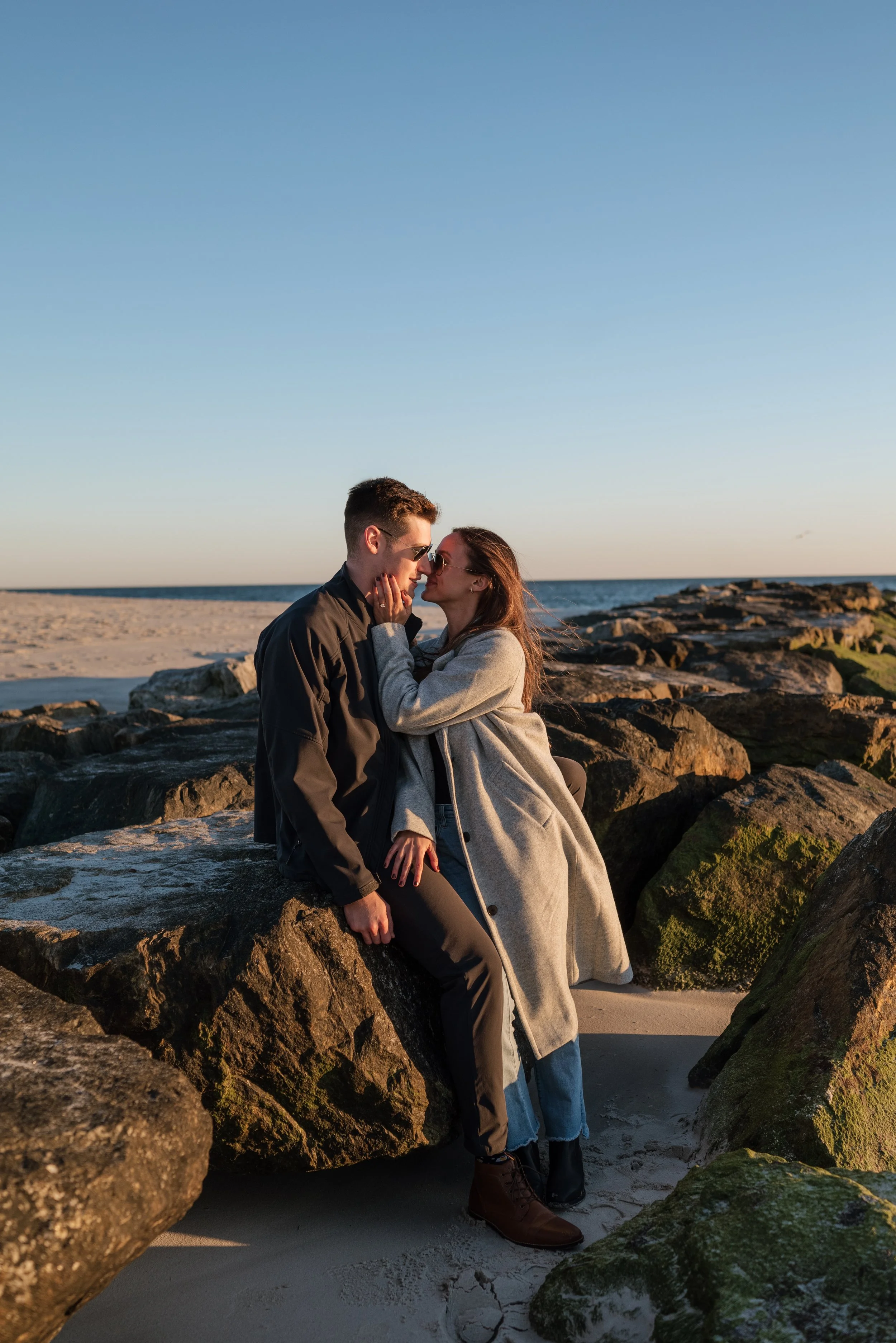 A couple sitting on rocks at the beach during sunset, close together and looking into each other's eyes.