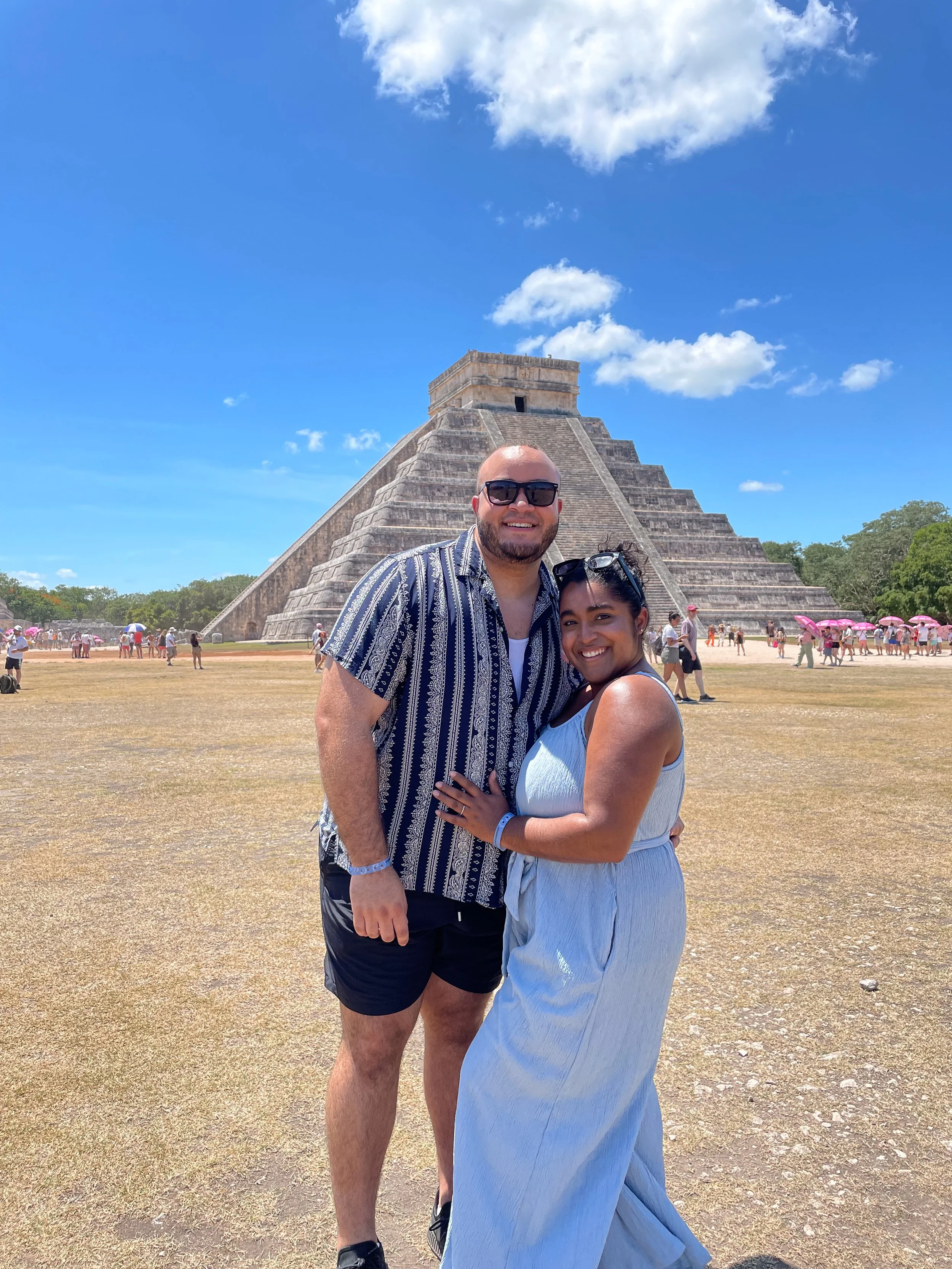 A smiling couple is standing in front of the ancient Mayan pyramid at Chichen Itza, with blue skies and some clouds overhead and other tourists around them.