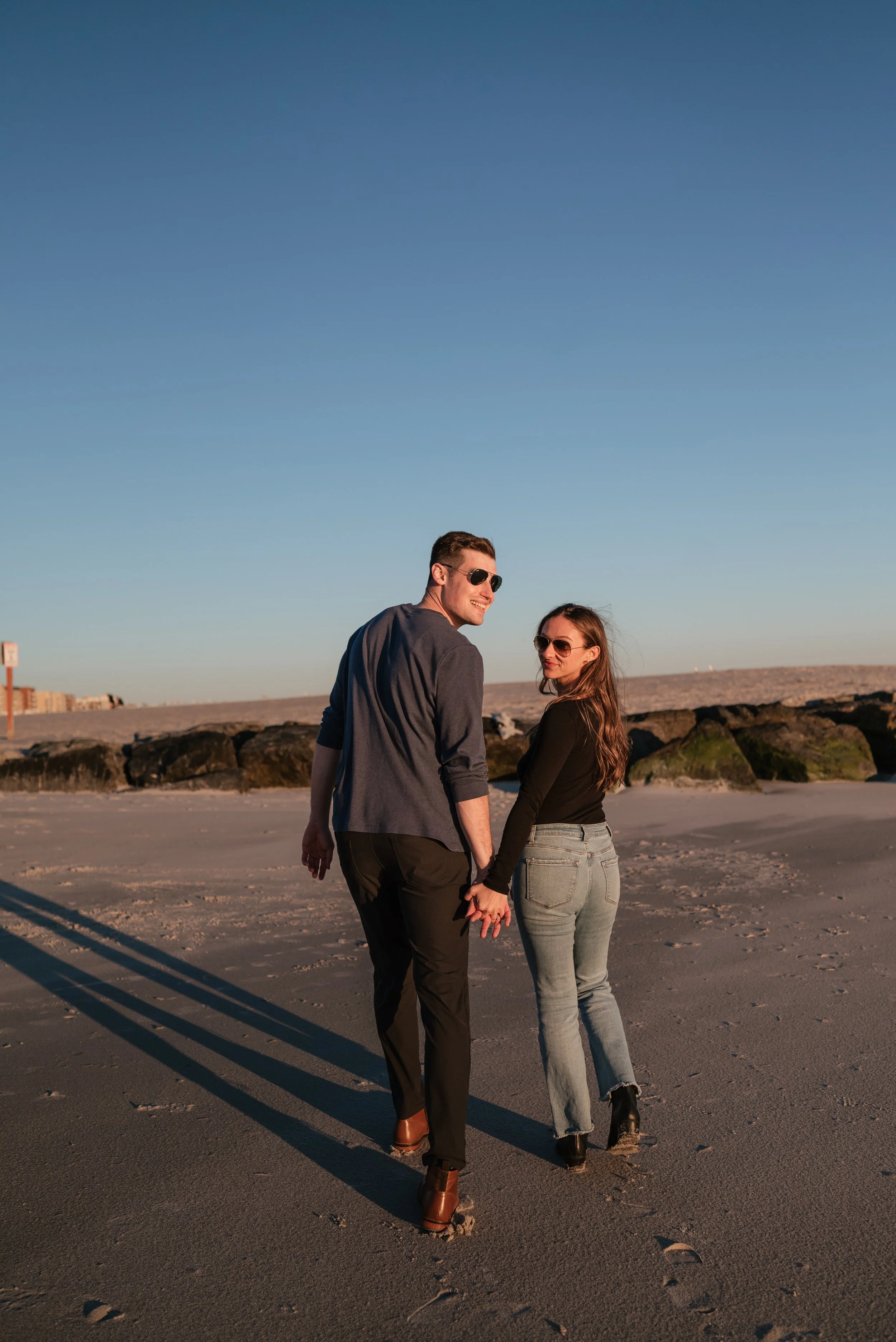 A young couple holding hands and walking on a beach at sunset, wearing sunglasses and casual clothing, with large rocks in the background and a clear blue sky.