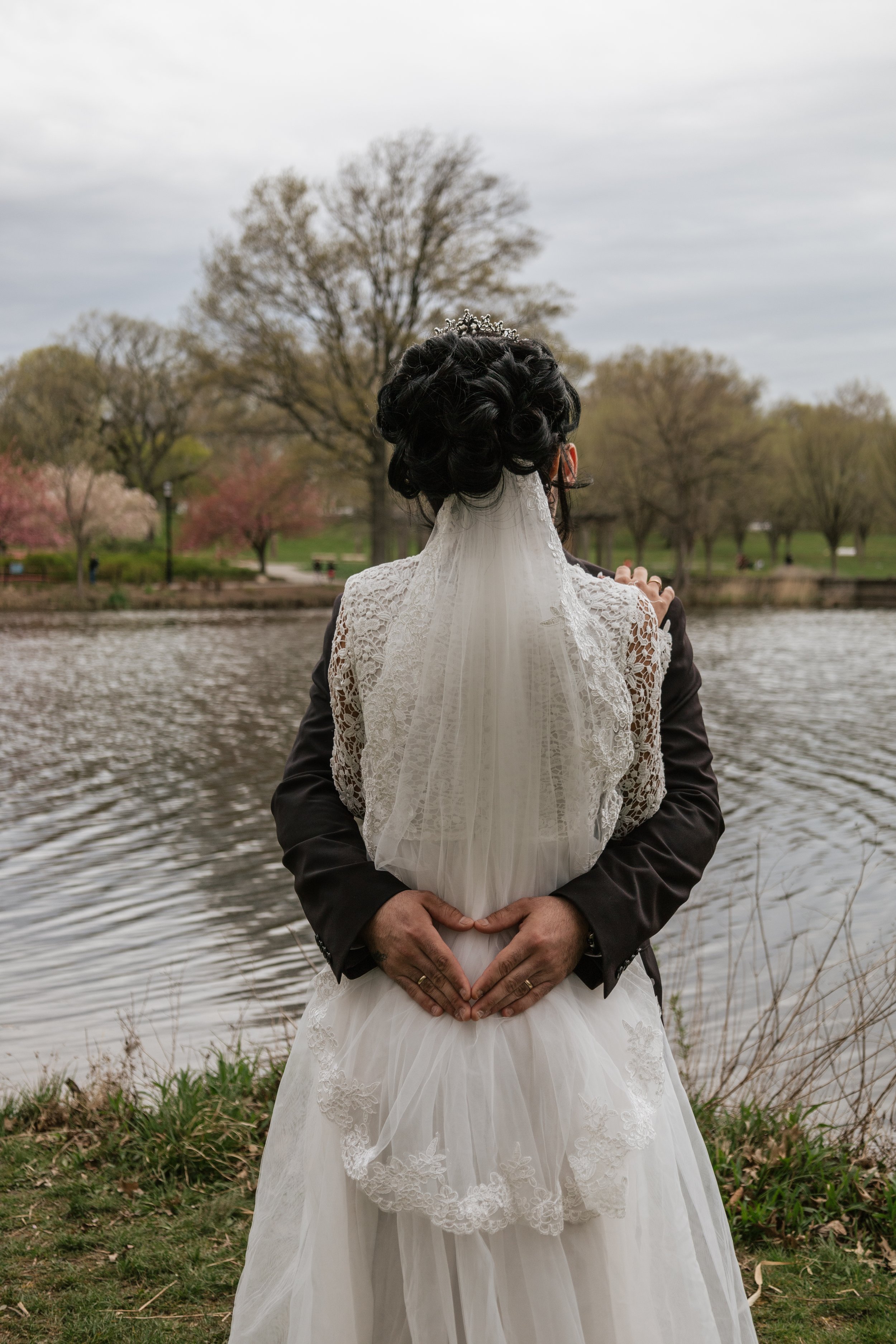 A bride and groom standing by a lake, with the groom embracing the bride from behind during an outdoor wedding in a park.