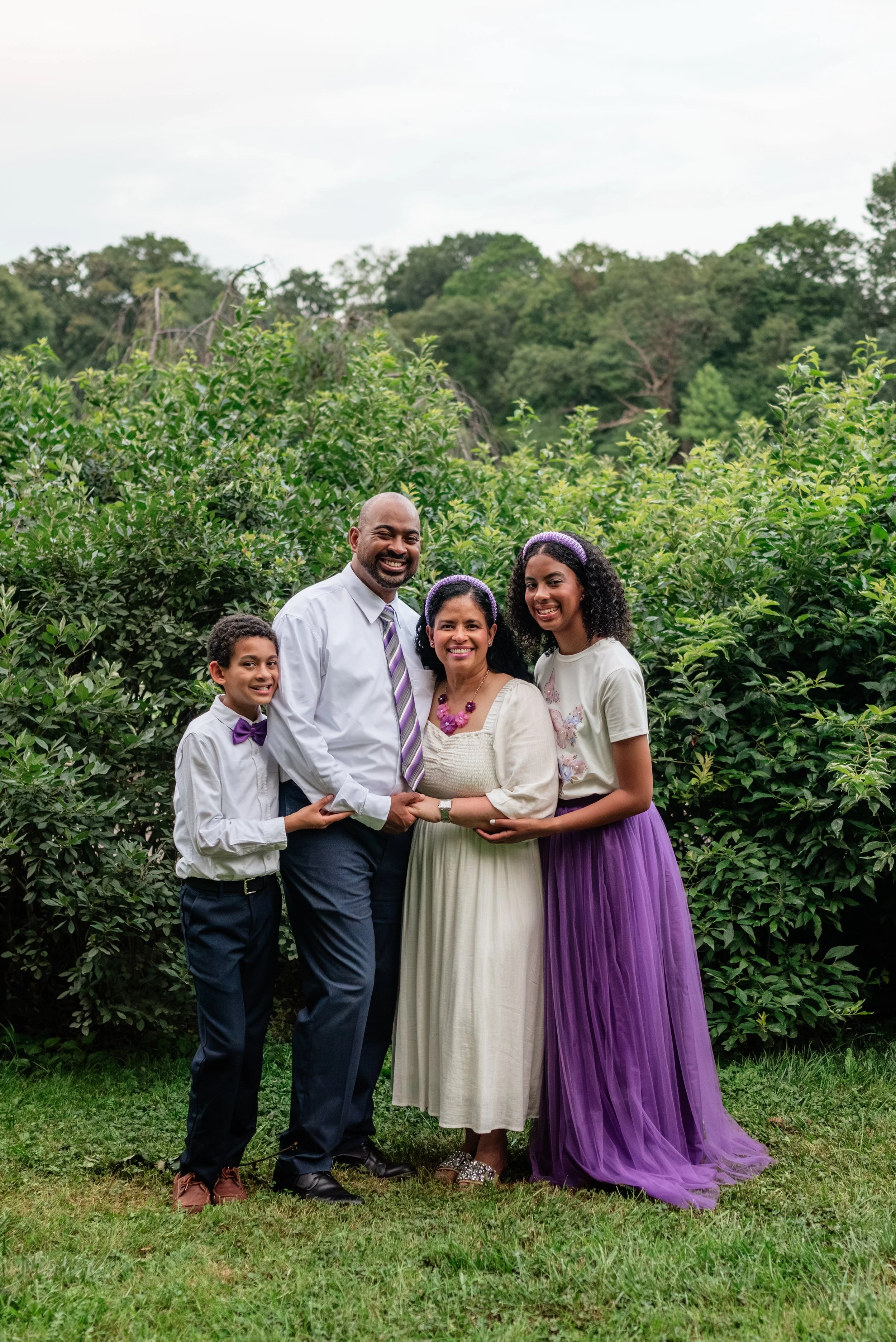 Family of four standing outdoors in front of lush greenery, smiling and holding hands, with a backdrop of trees and cloudy sky.