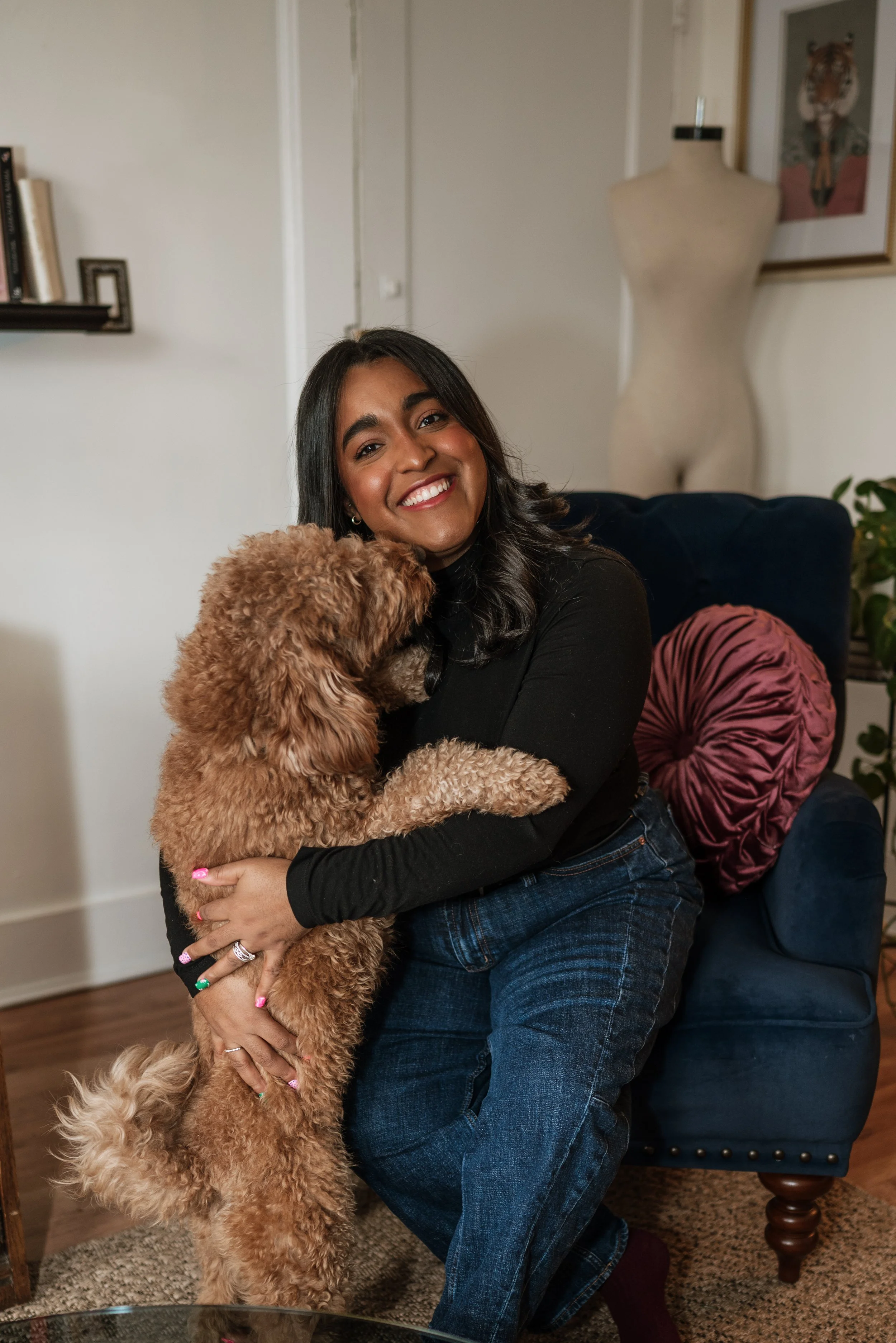 A woman with dark hair, wearing a black top, is hugging a fluffy brown dog in a living room.