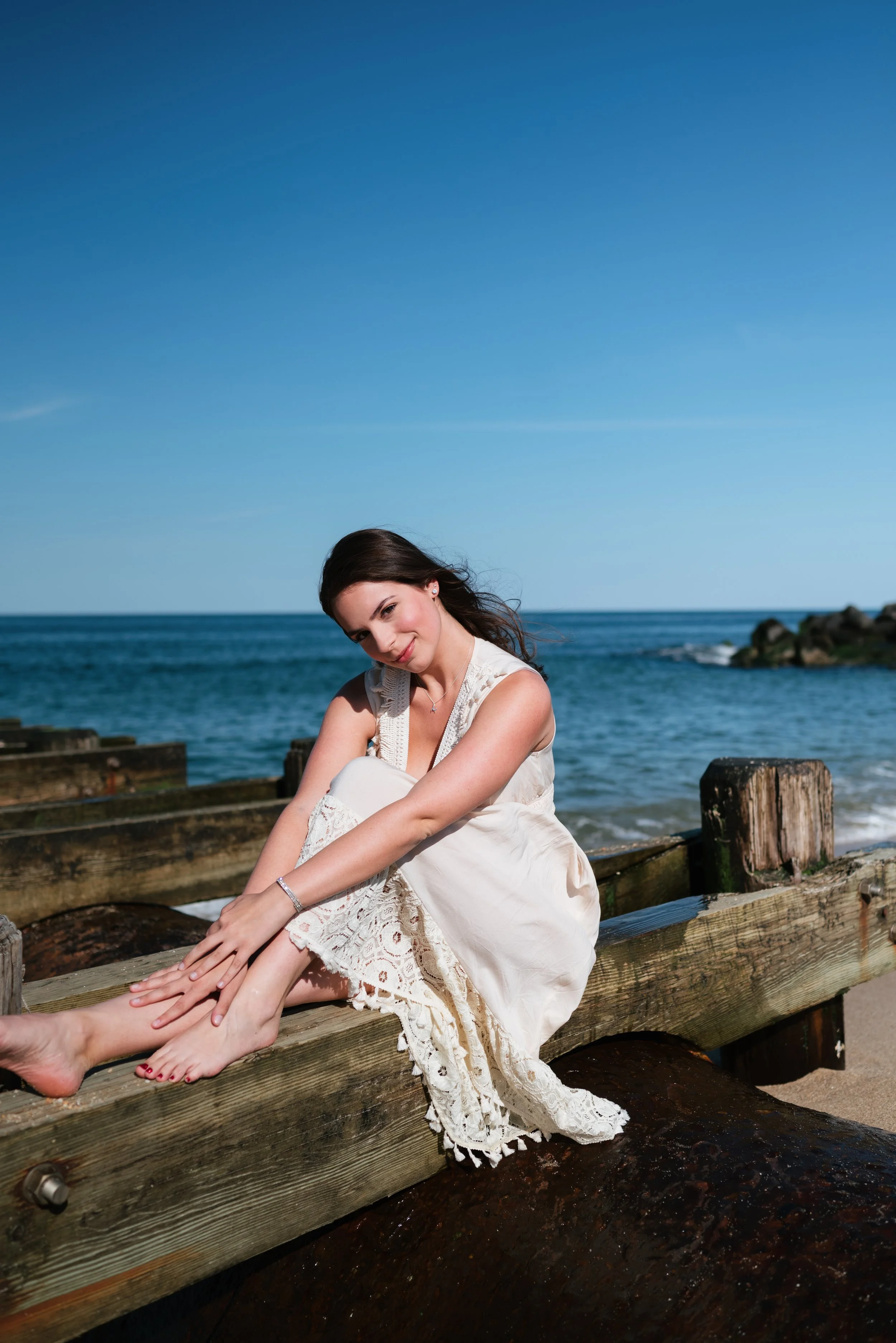 A woman sitting on a wooden pier by the ocean, wearing a white dress, with dark hair, and a slight smile, under a clear blue sky.