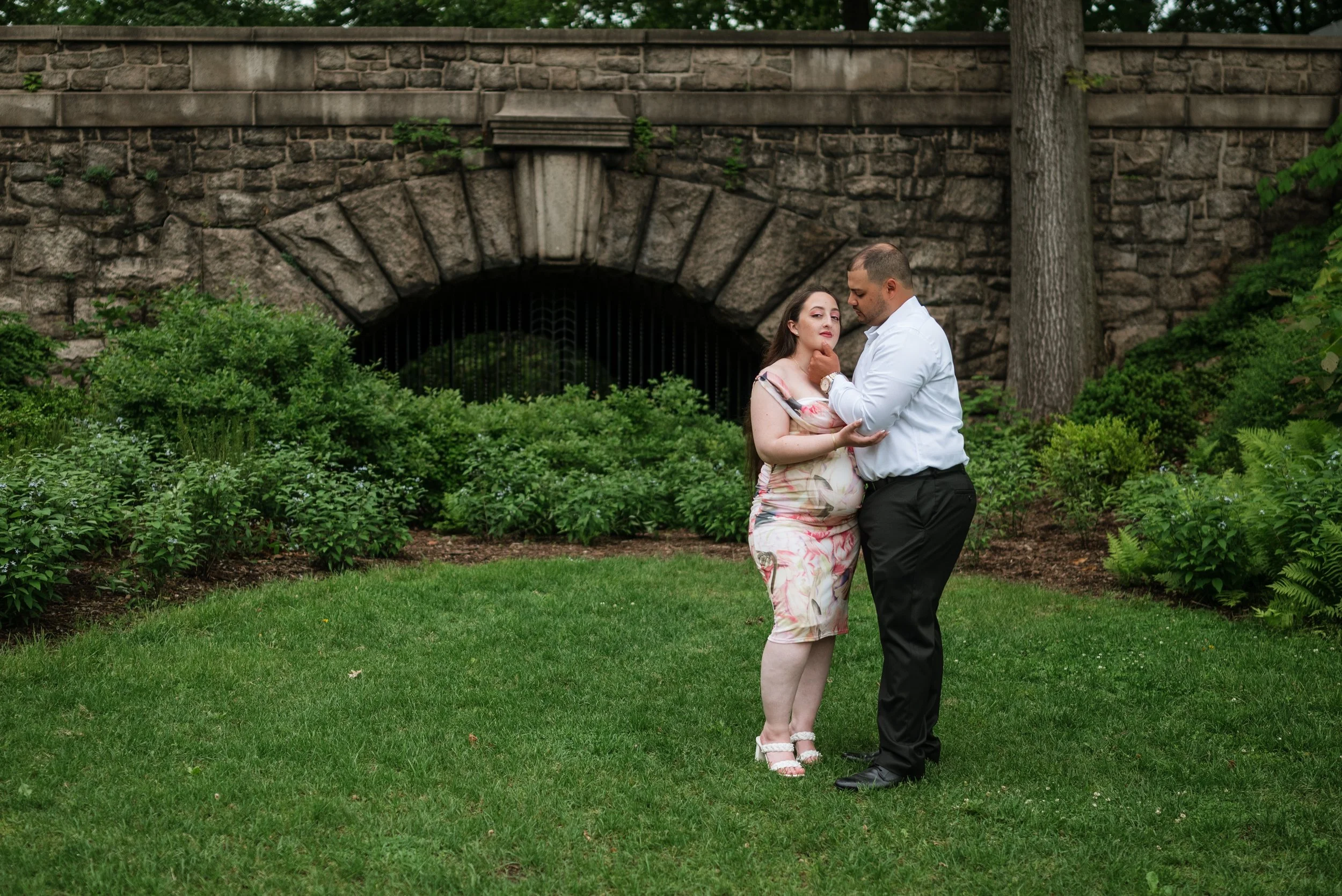 A pregnant woman and a man stand close together on a grassy area, holding each other in front of a brick arched bridge with greenery surrounding them.