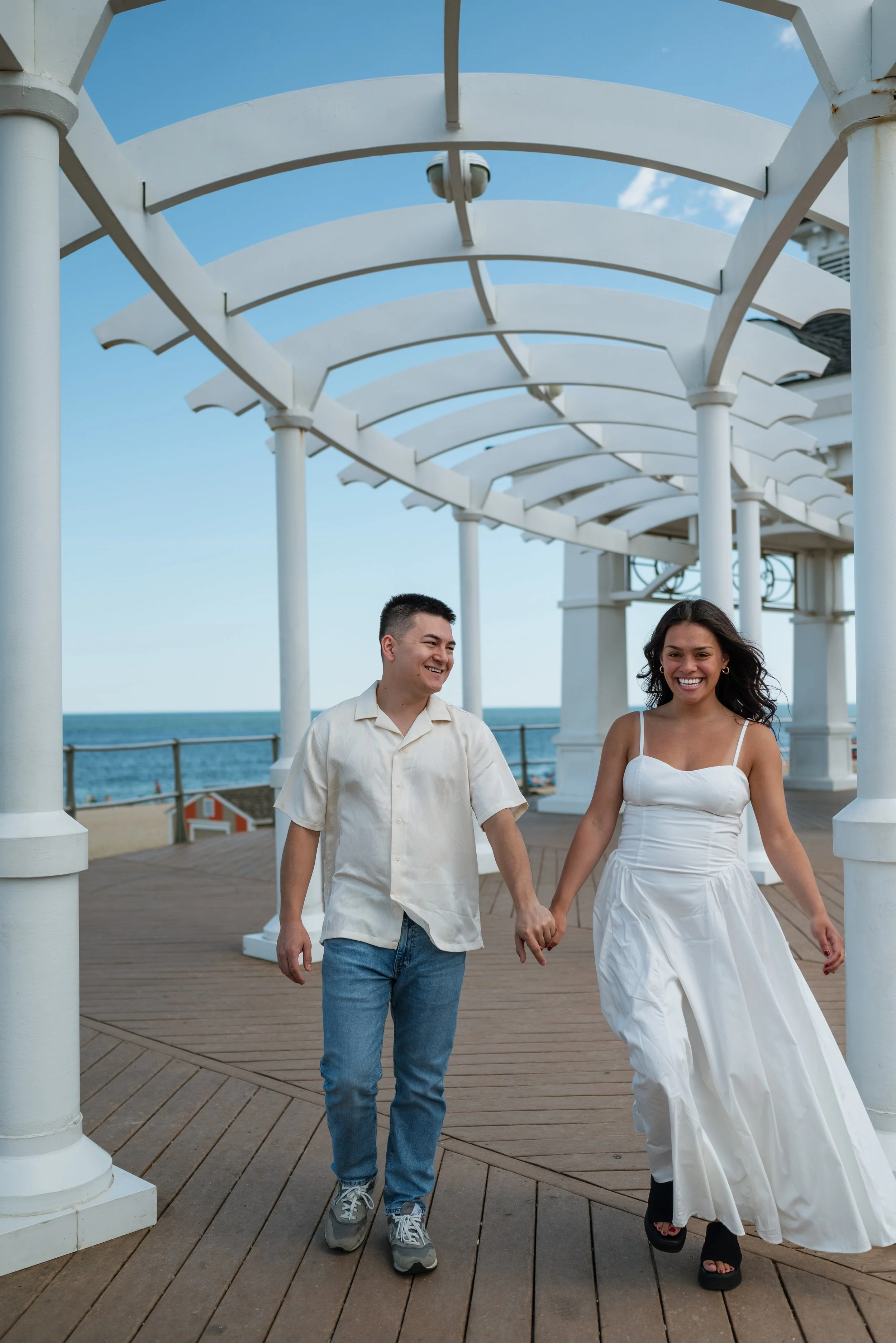 A smiling couple walking hand in hand on a boardwalk near the ocean under a white pergola, with clear blue sky and some clouds in the background.