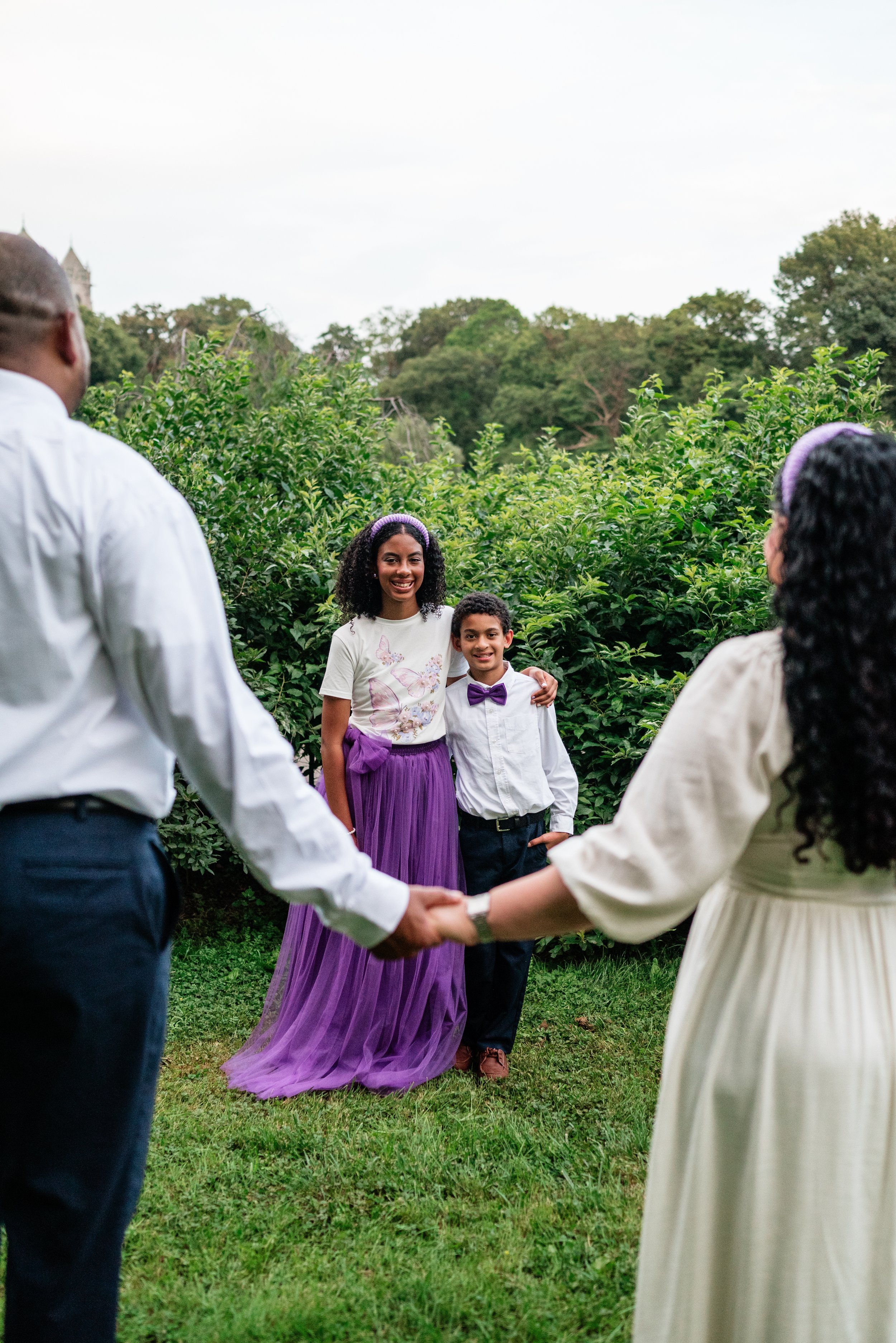 A young woman and a boy stand together smiling in front of green shrubs, while holding hands with a couple in the foreground who are holding hands.