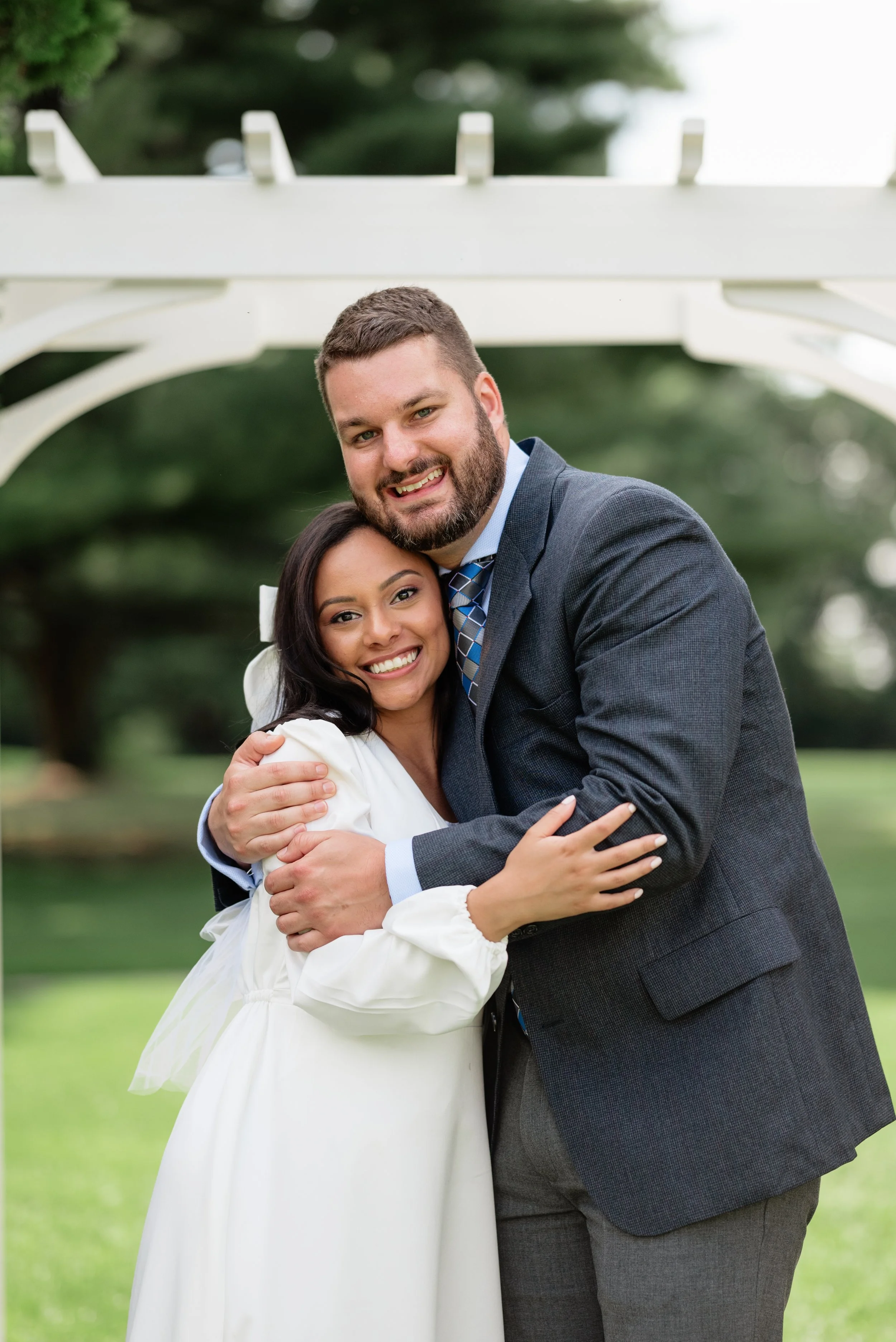 A smiling couple hugging outdoors in a park, with the woman wearing a white dress and the man in a gray suit, under a white outdoor arch.