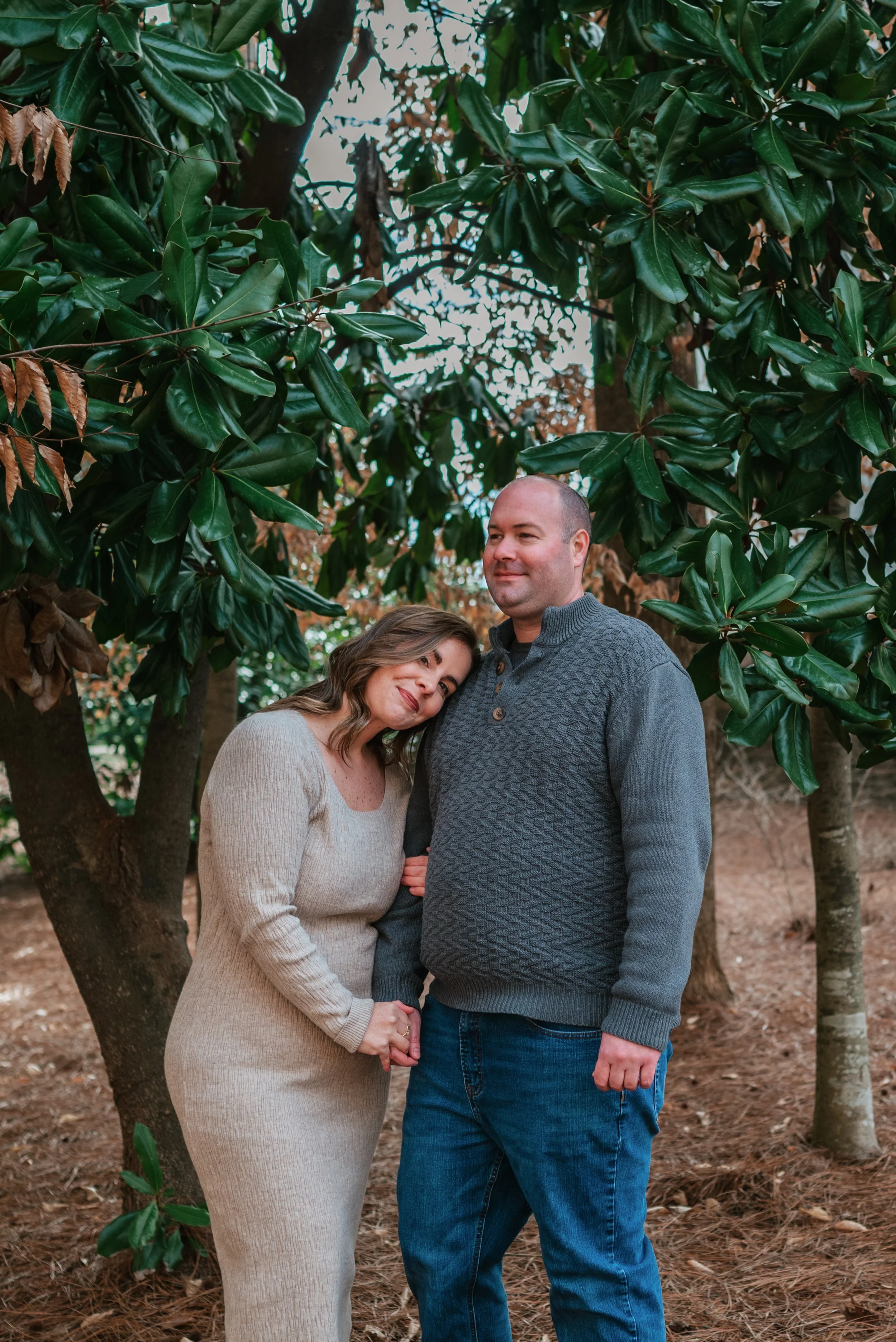 A couple standing outdoors among trees, with the woman resting her head on the man's shoulder and holding his hand.