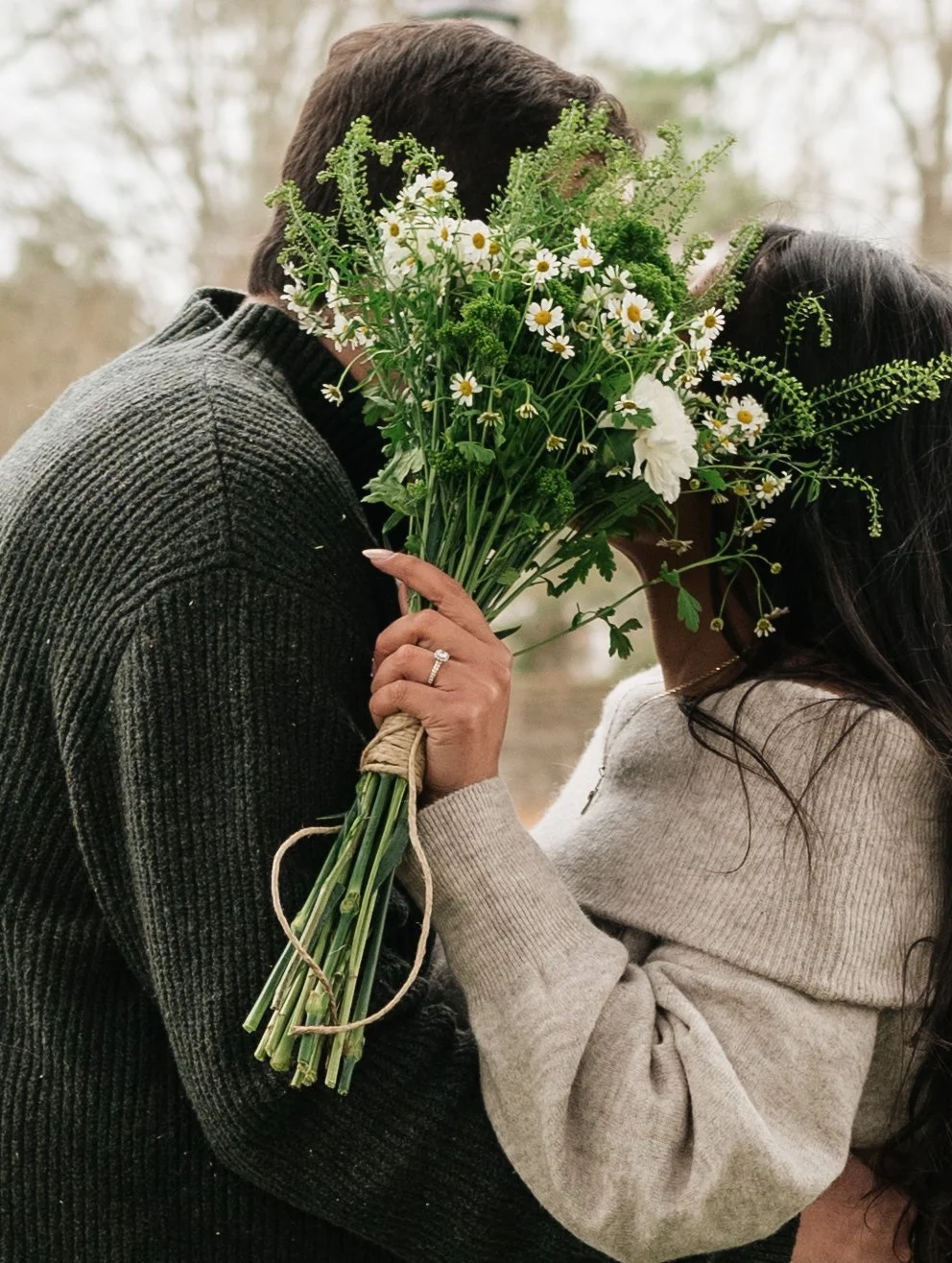 A couple holding a bouquet of white daisies and greenery, with their faces close together in an outdoor setting.