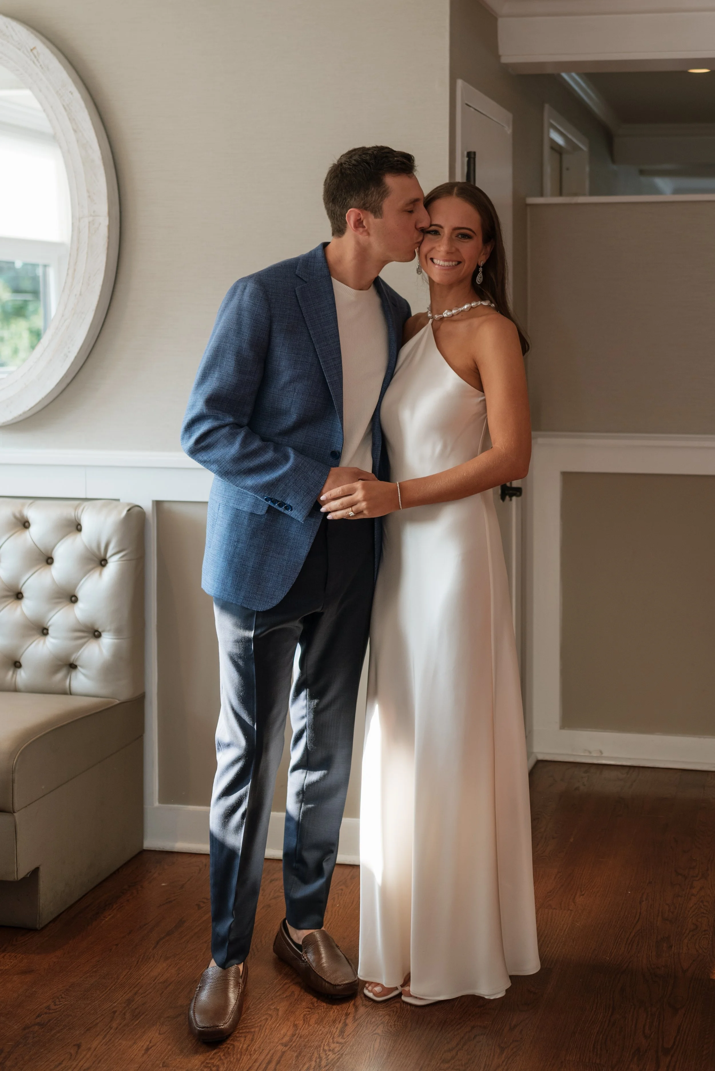 A couple at a wedding, the man in a blue blazer and grey pants kissing the woman's temple, who is in a white satin gown smiling.