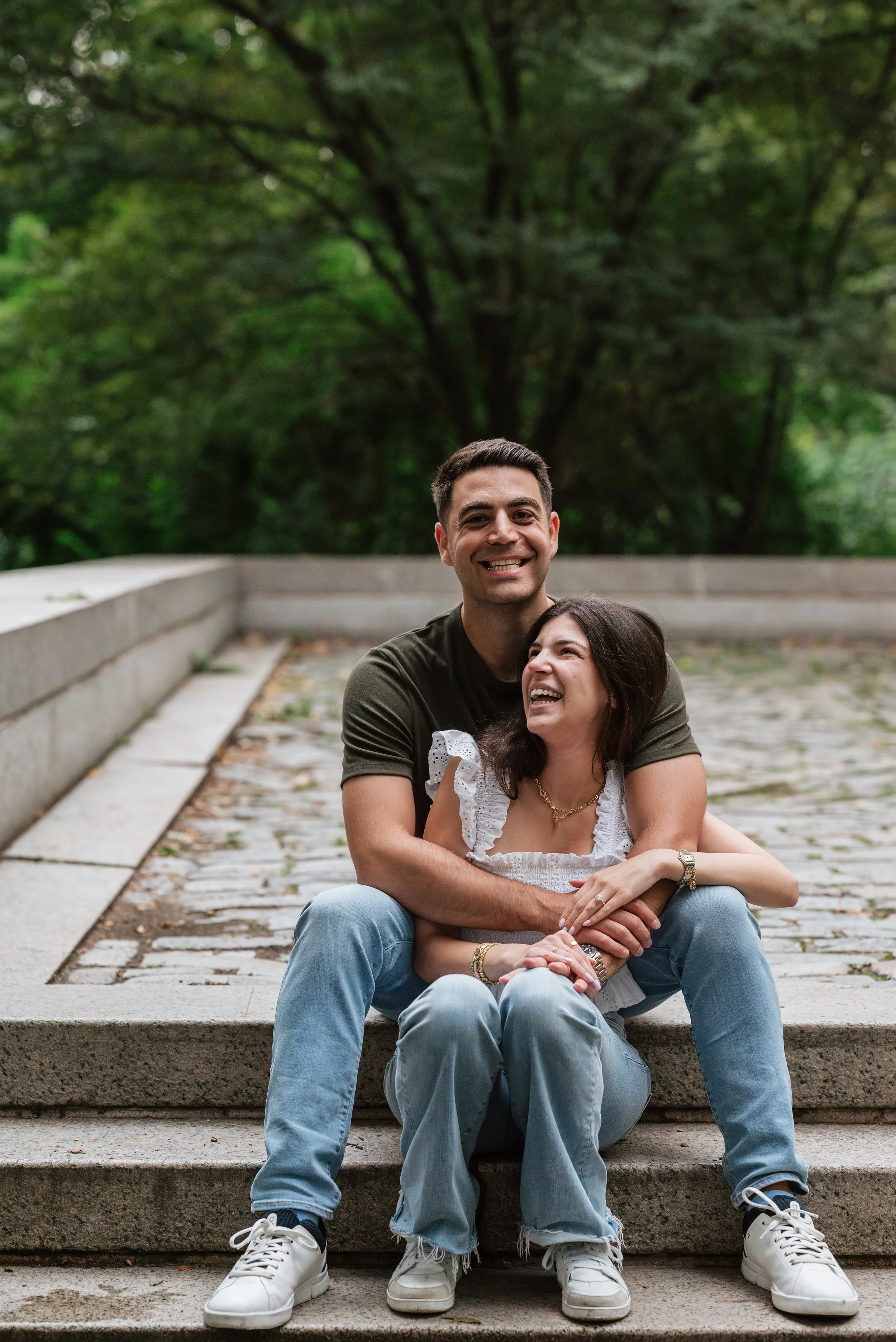 A happy couple sitting on outdoor steps in a park, smiling and embracing each other, surrounded by green trees.