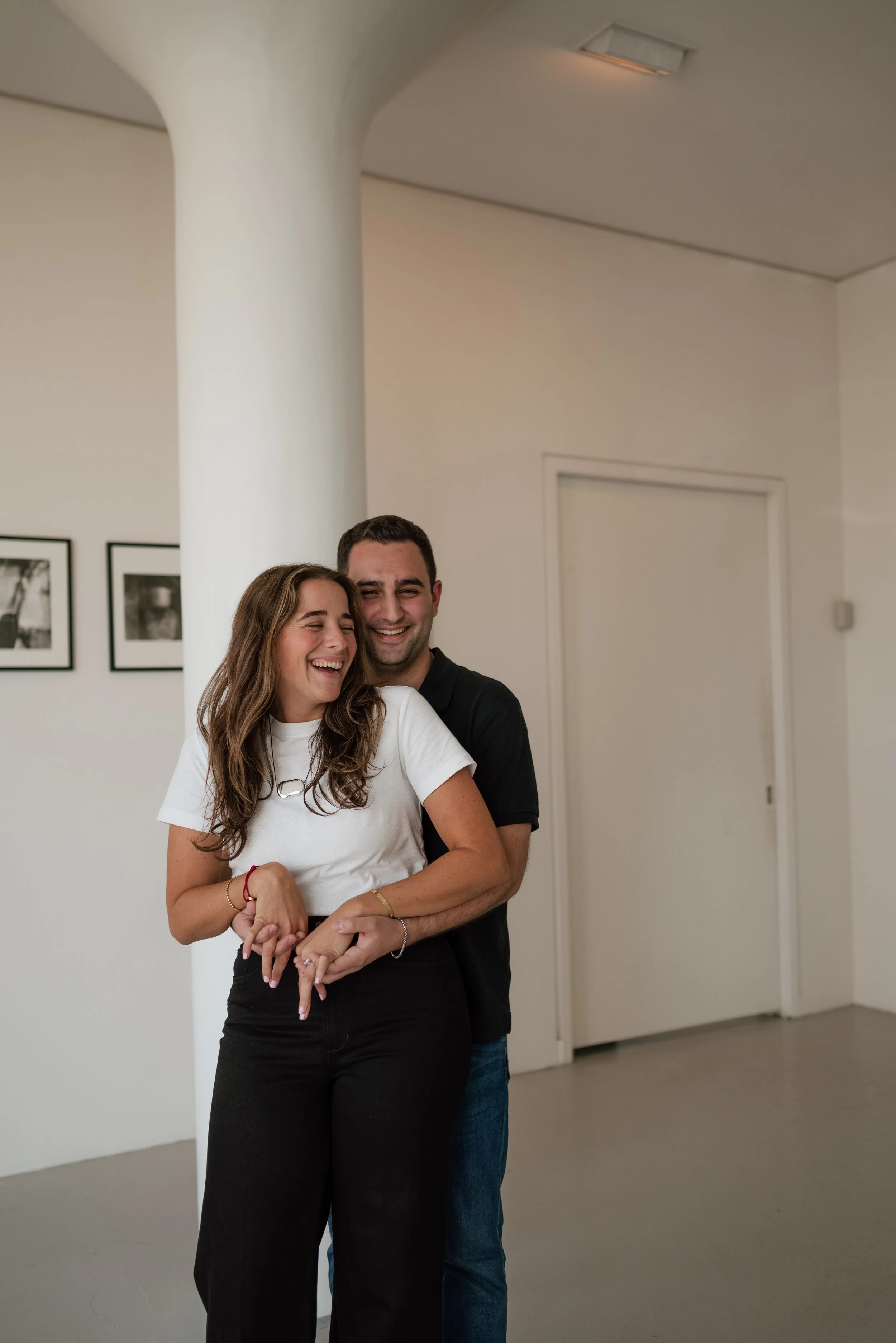 Two people laughing and hugging each other in an indoor space with framed photographs on the wall.