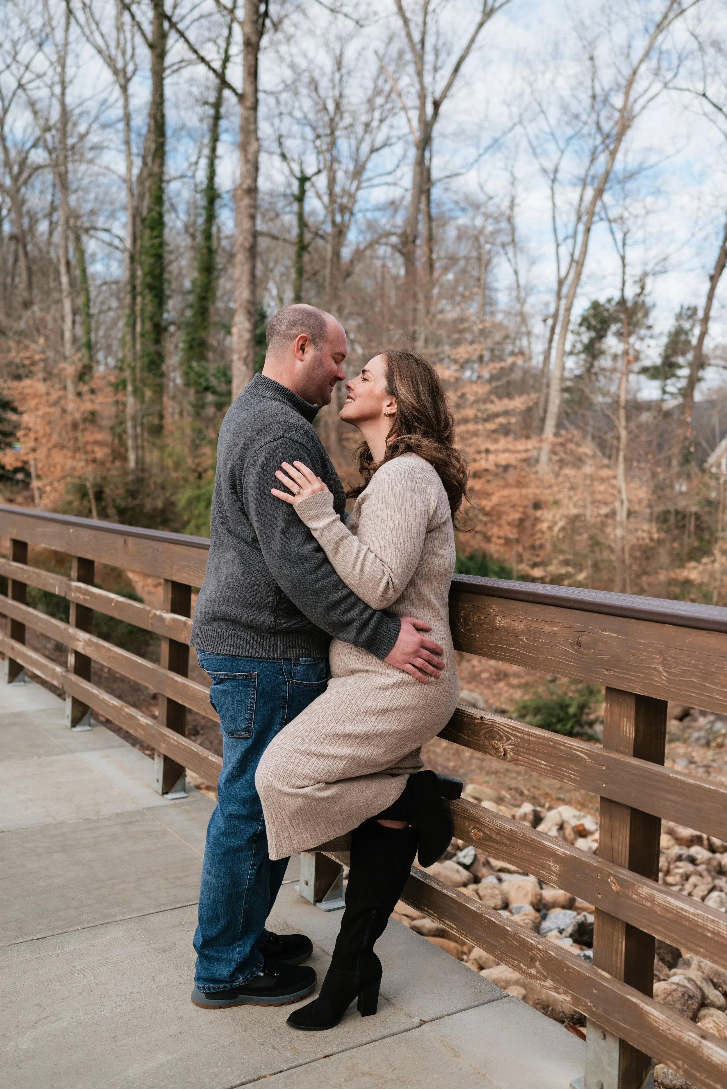 A couple sharing a romantic moment on a wooden bridge outdoors, with trees and fall foliage in the background.