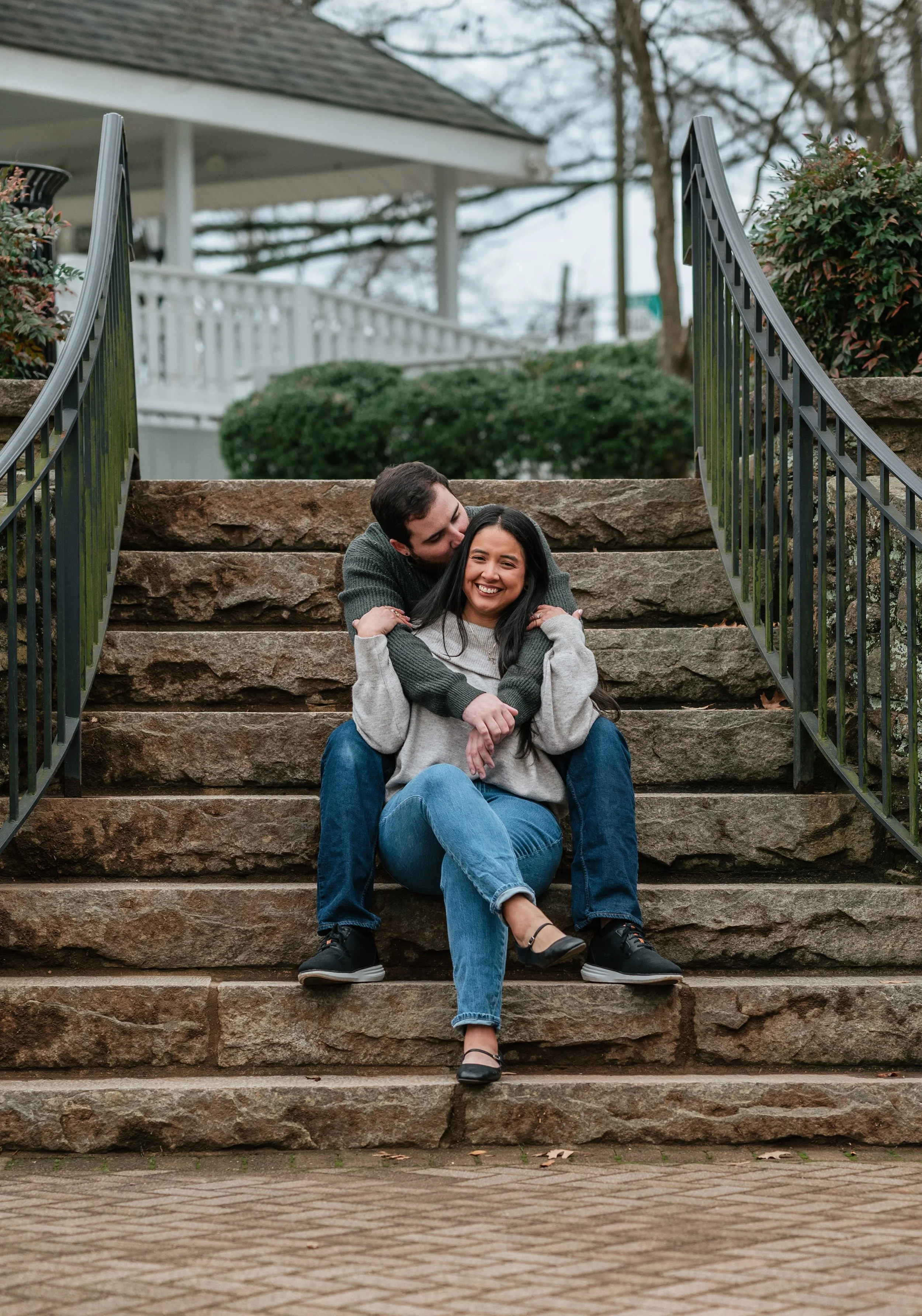 A couple sitting on stone stairs outdoors, embracing and smiling at each other on a cloudy day.