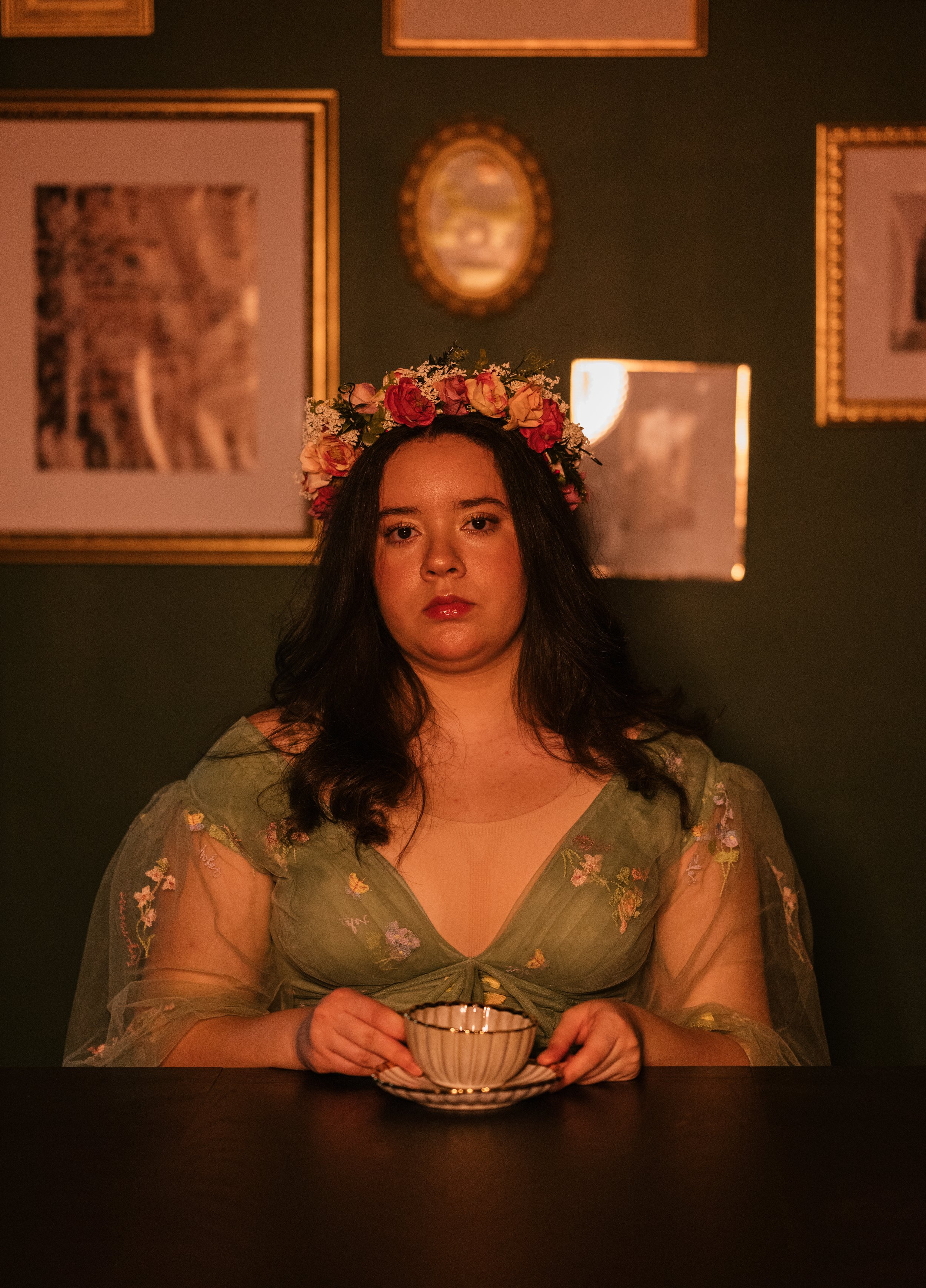 A woman with dark hair wearing a floral crown and a green embroidered dress with sheer sleeves, sitting at a dark table holding a teacup and saucer. The background features framed pictures on a dark wall and warm lighting.