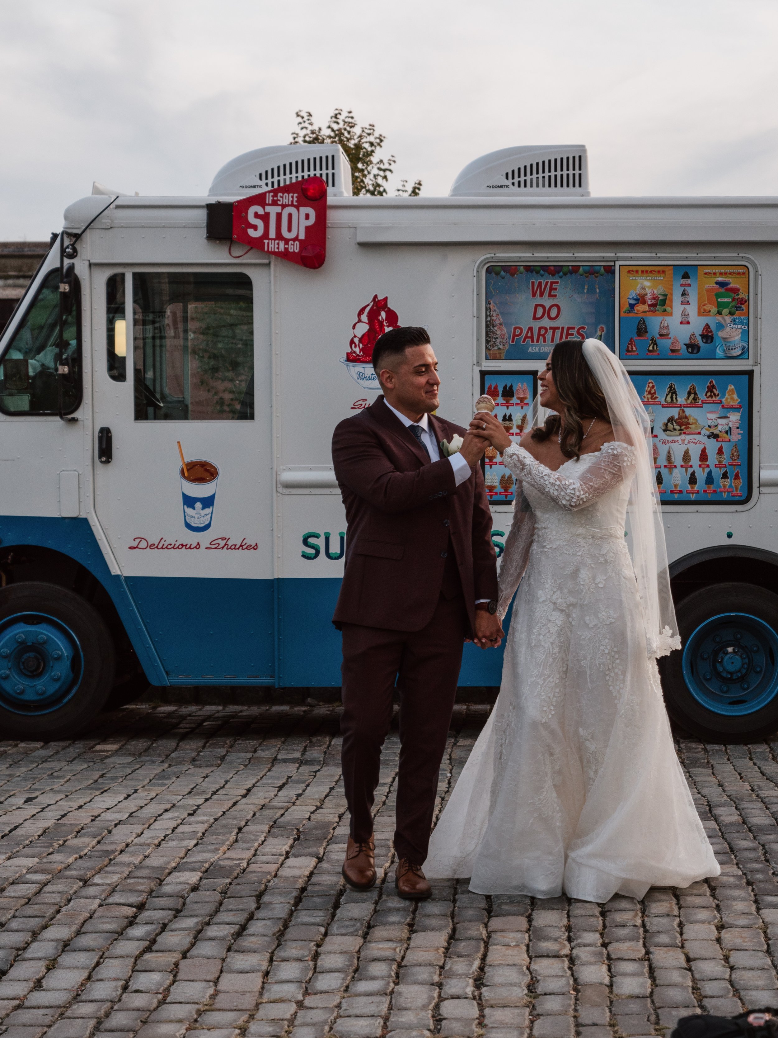 A newlywed couple in wedding attire sharing a moment in front of an ice cream truck, on a cobblestone street.