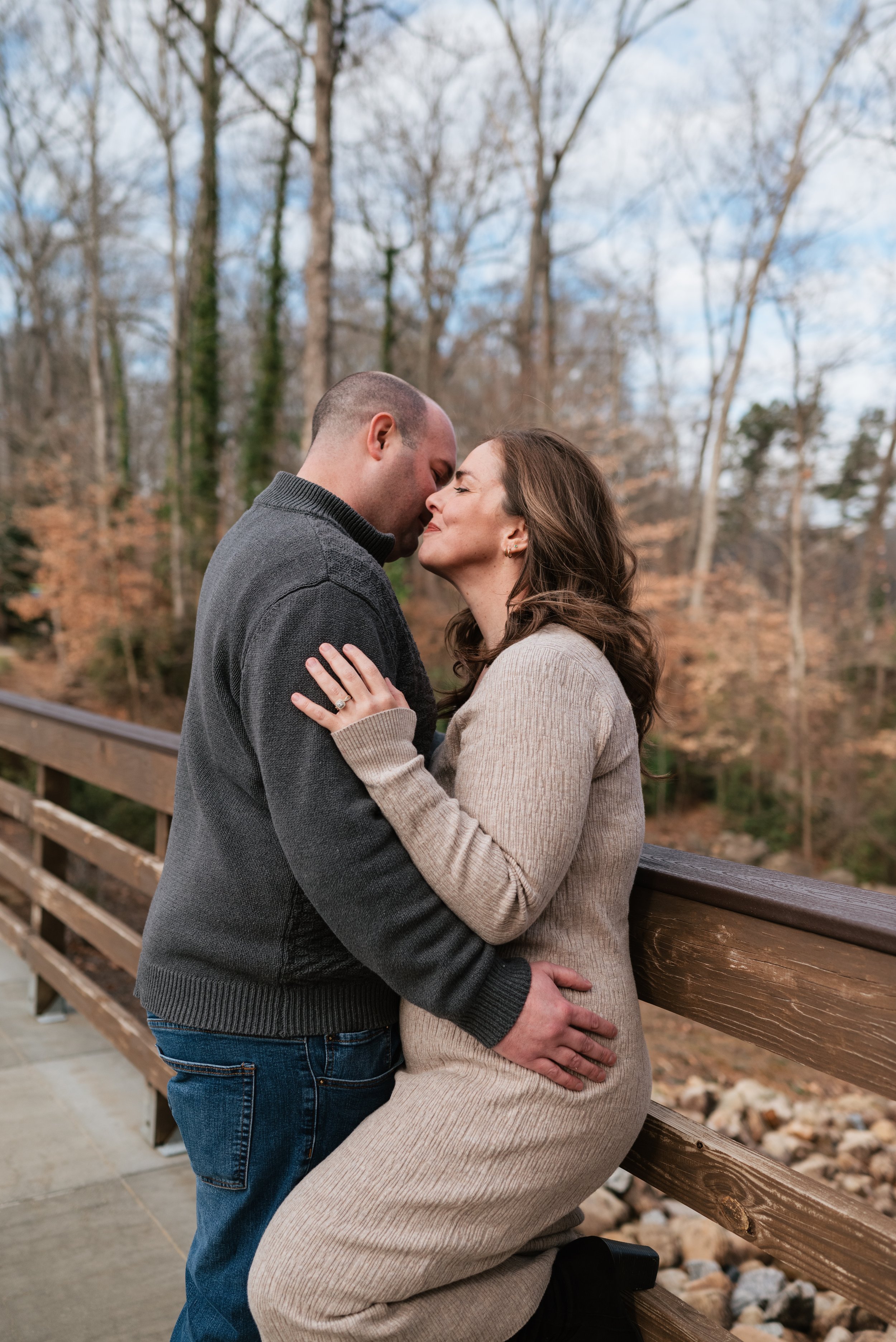 A couple sharing a tender moment in their engagement photoshoot in downtown Alpharetta, Georgia with foreheads touching, standing on a wooden bridge with trees in the background.