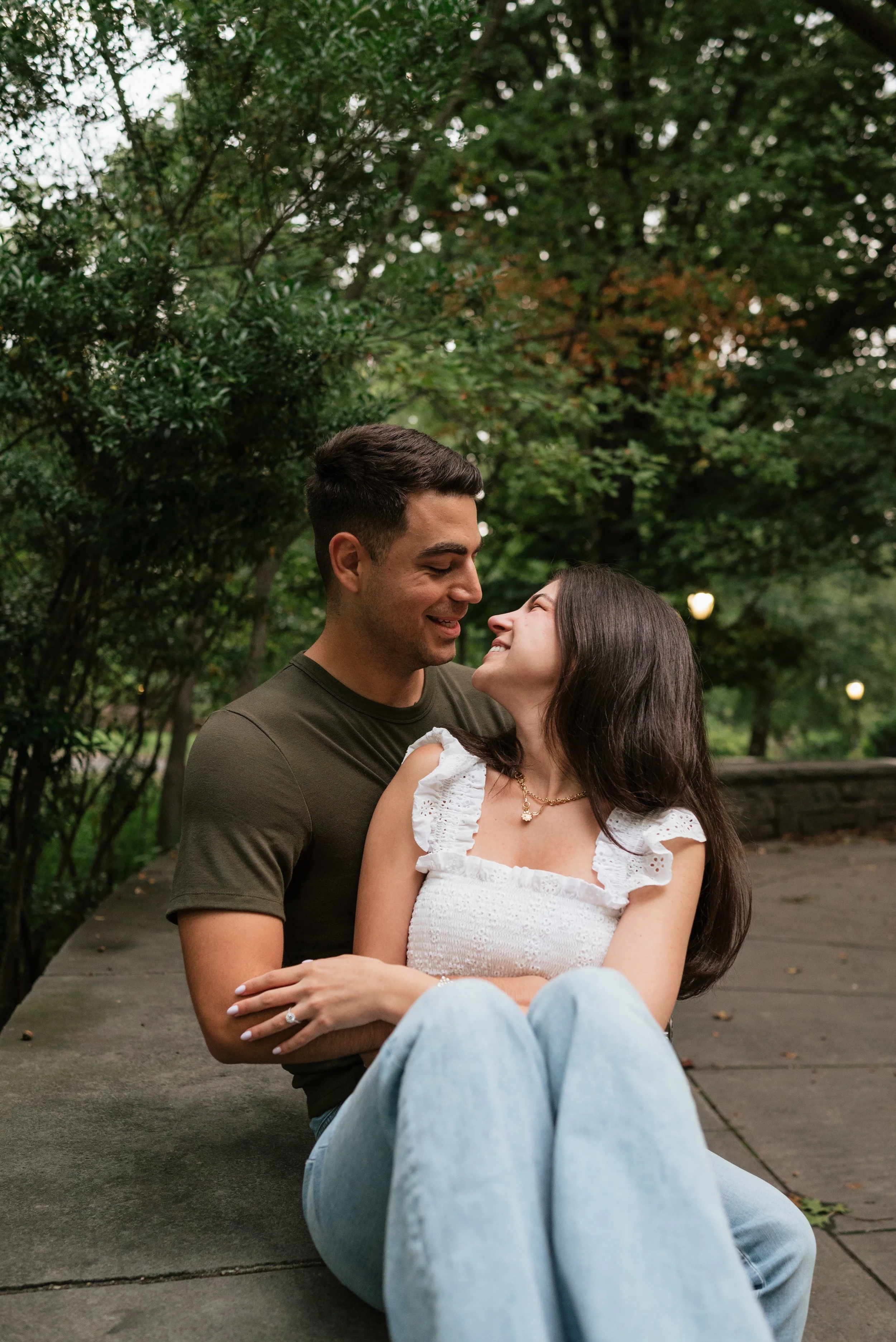 A couple sitting on a concrete path in a park, looking at each other and smiling, surrounded by greenery.