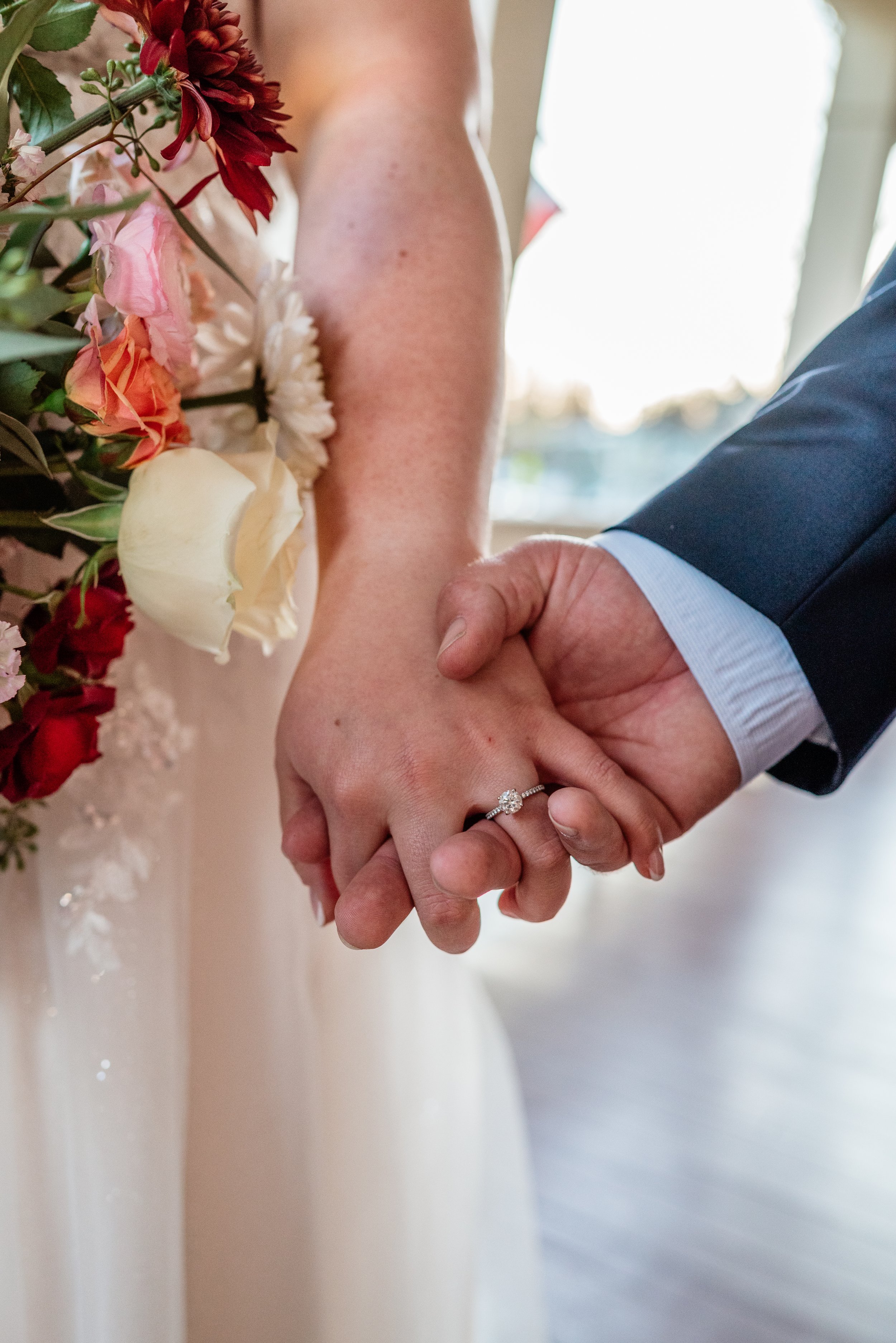 Close-up of a bride and groom holding hands during a wedding, with the bride wearing a diamond engagement ring, and the bride holding a bouquet of flowers.