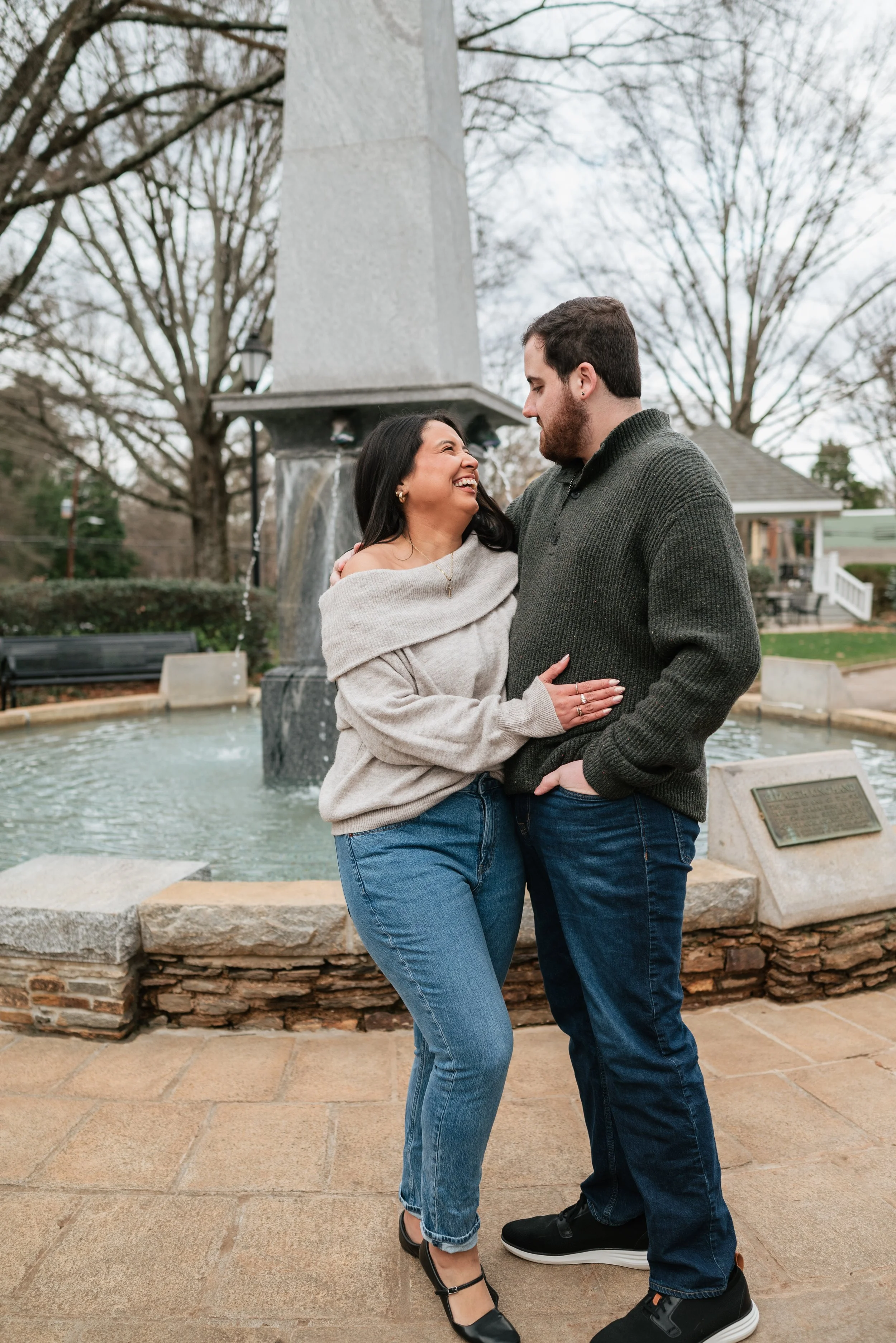 A couple standing close and smiling at each other in front of a fountain in a park during winter.