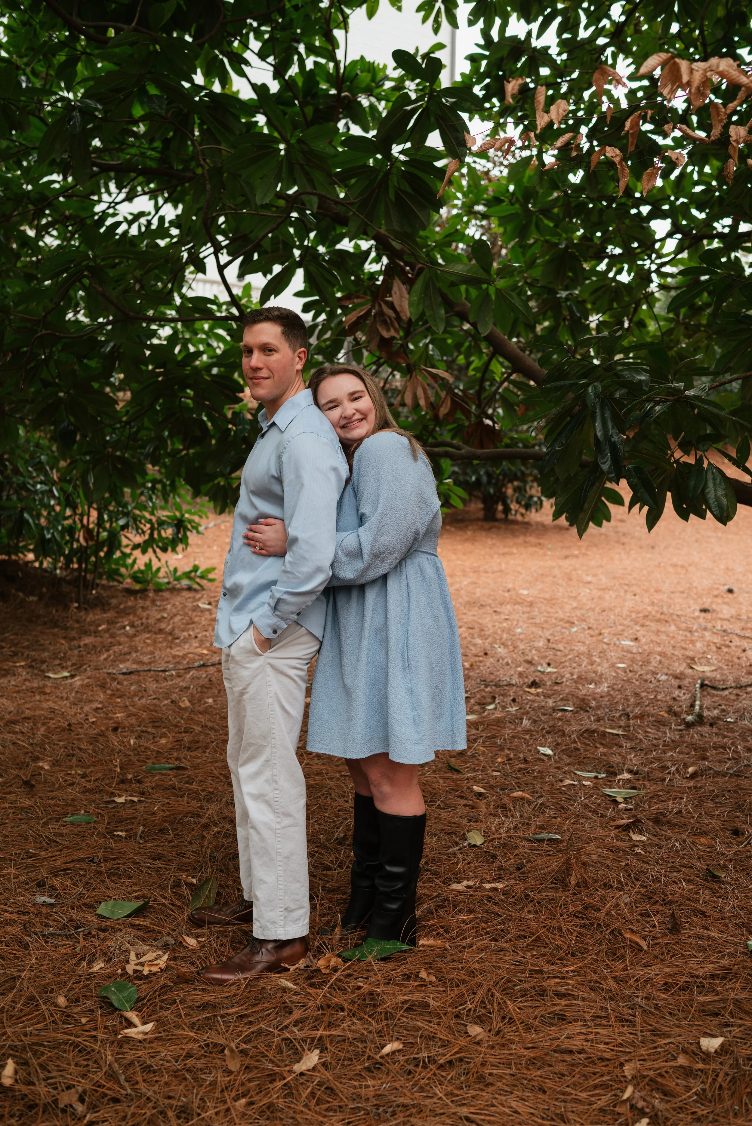 A young couple standing in a wooded area with trees and fallen leaves, smiling and hugging, dressed in light blue and neutral-colored clothing.