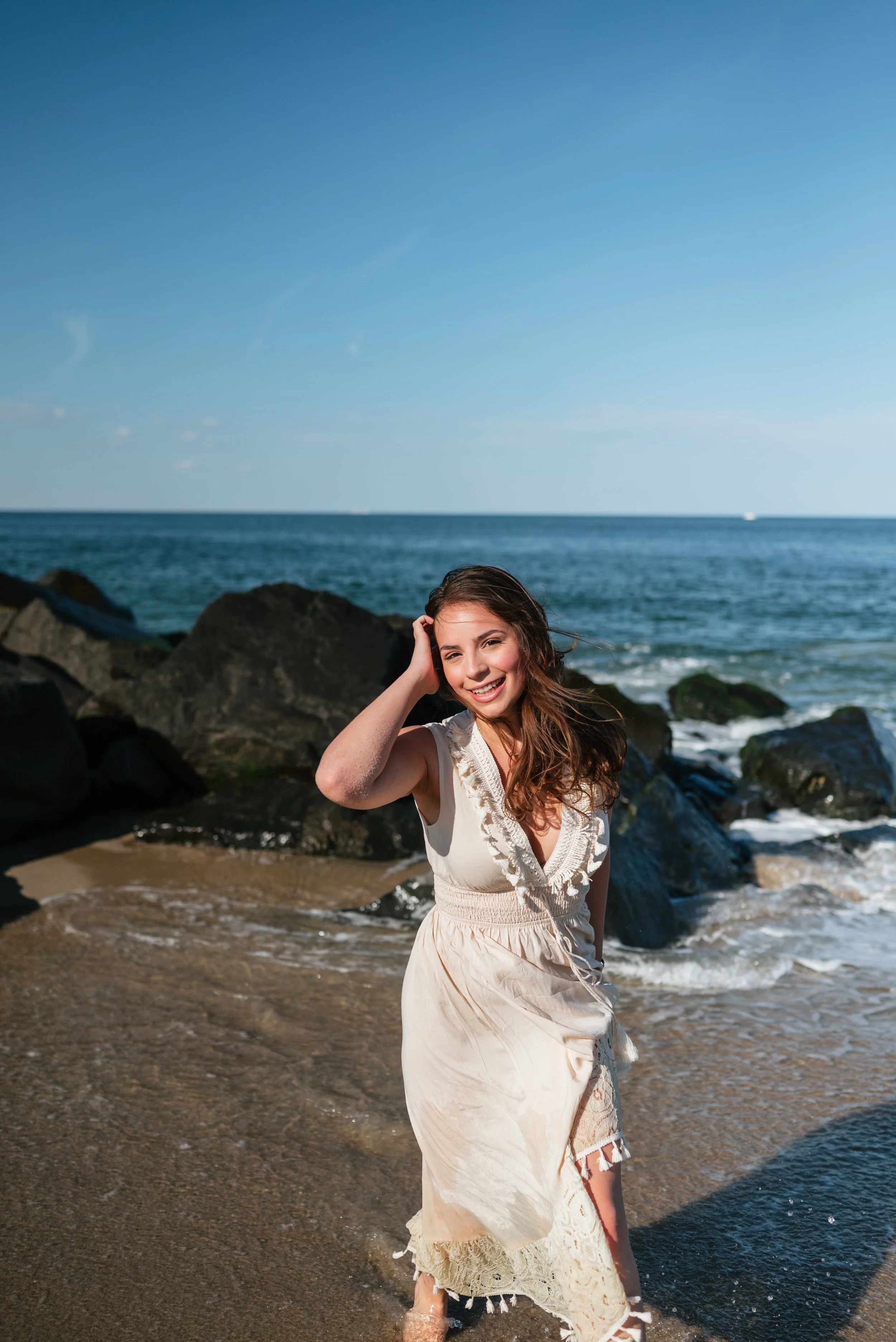 A woman with long brown hair in a white dress standing on a sandy beach near rocks with the ocean and blue sky in the background.