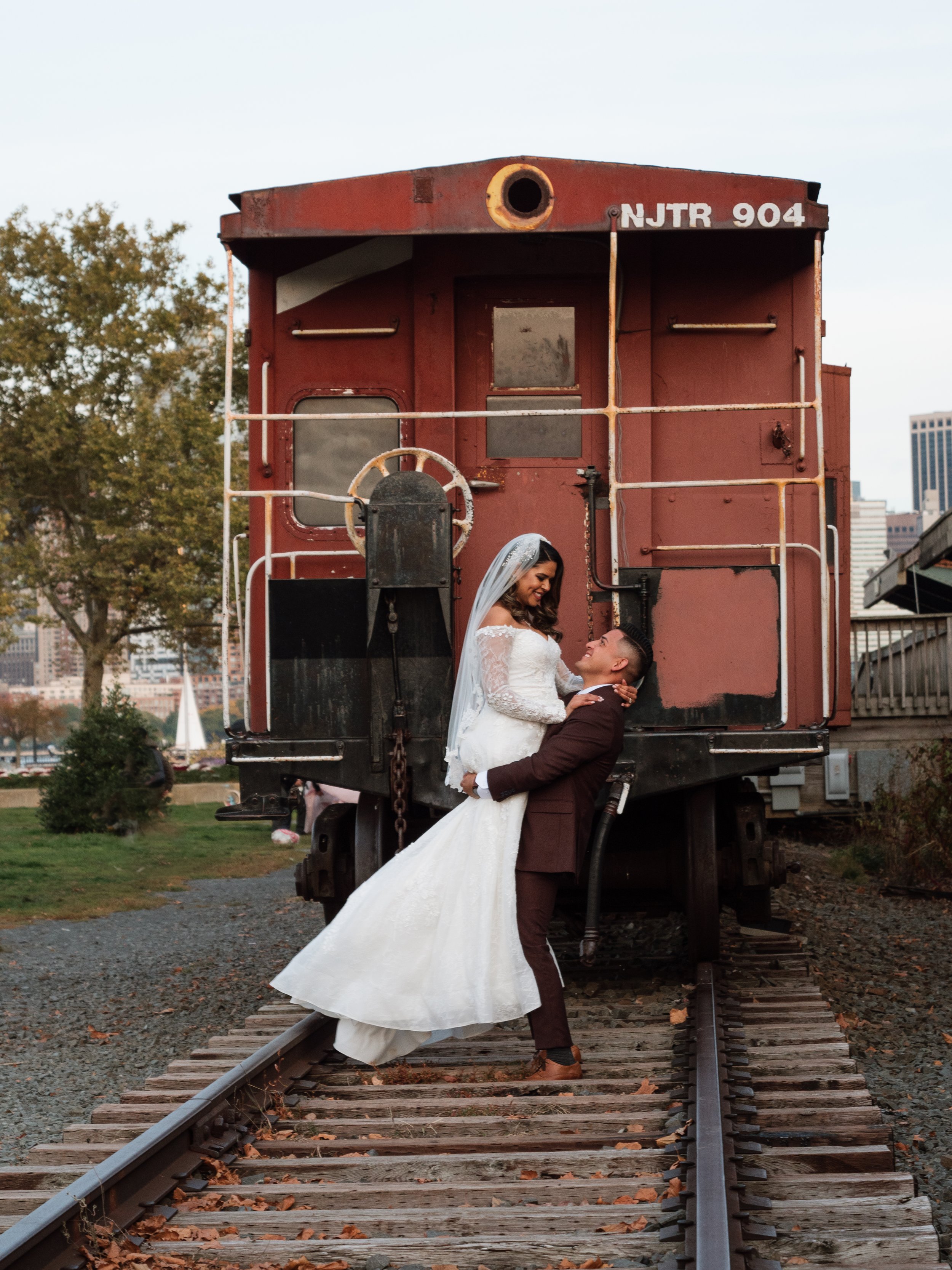 A bride and groom embrace in front of a vintage train car on railroad tracks, with city skyline in the background.