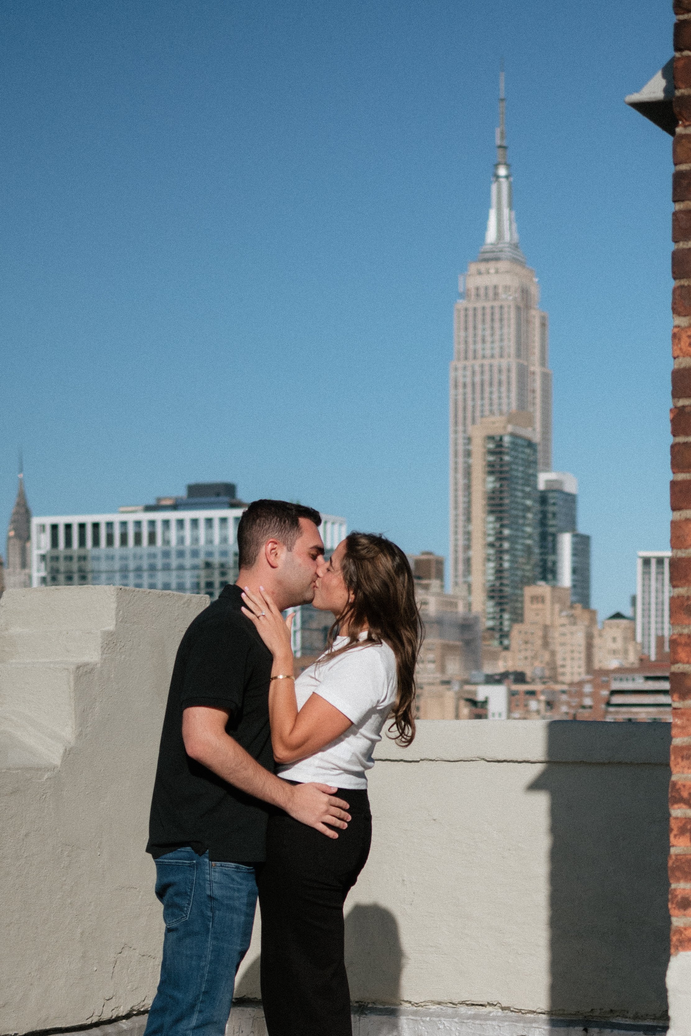Couple kissing on rooftop with New York City skyline, including Empire State Building, in the background on a sunny day.