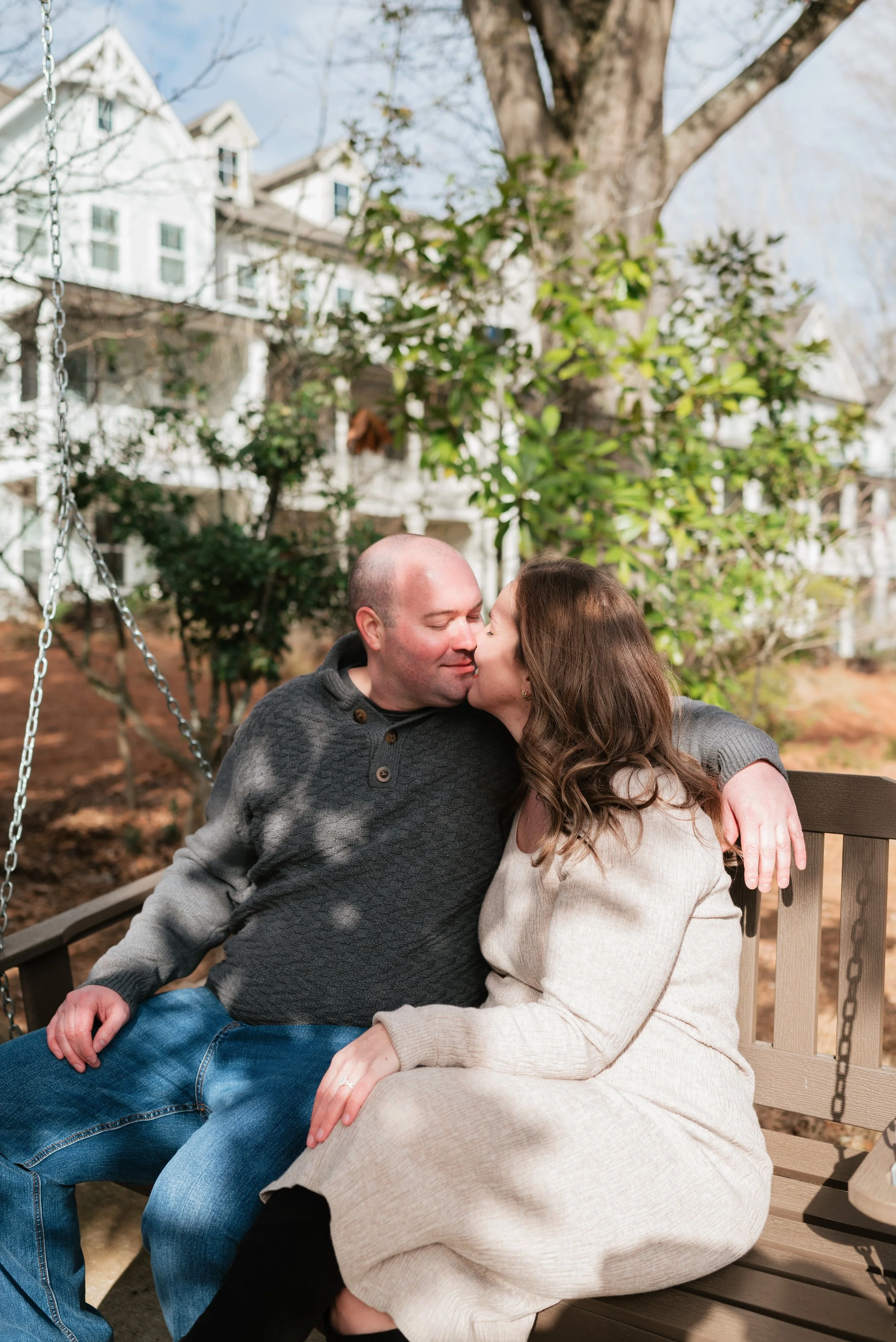 A couple sitting on a wooden park bench sharing a kiss outdoors with trees and houses in the background.