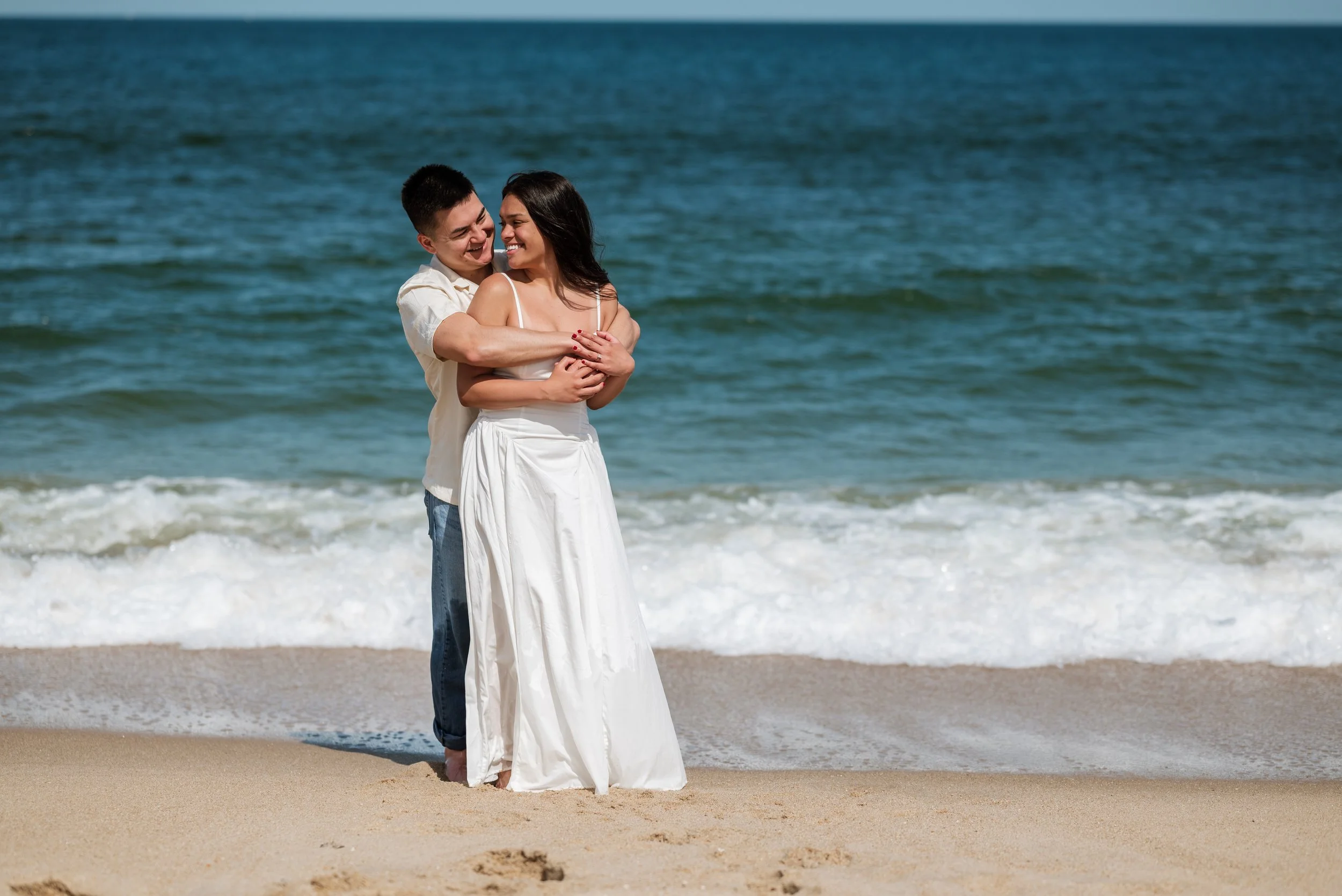 A smiling couple standing on the beach by the ocean, embracing each other with the waves in the background.