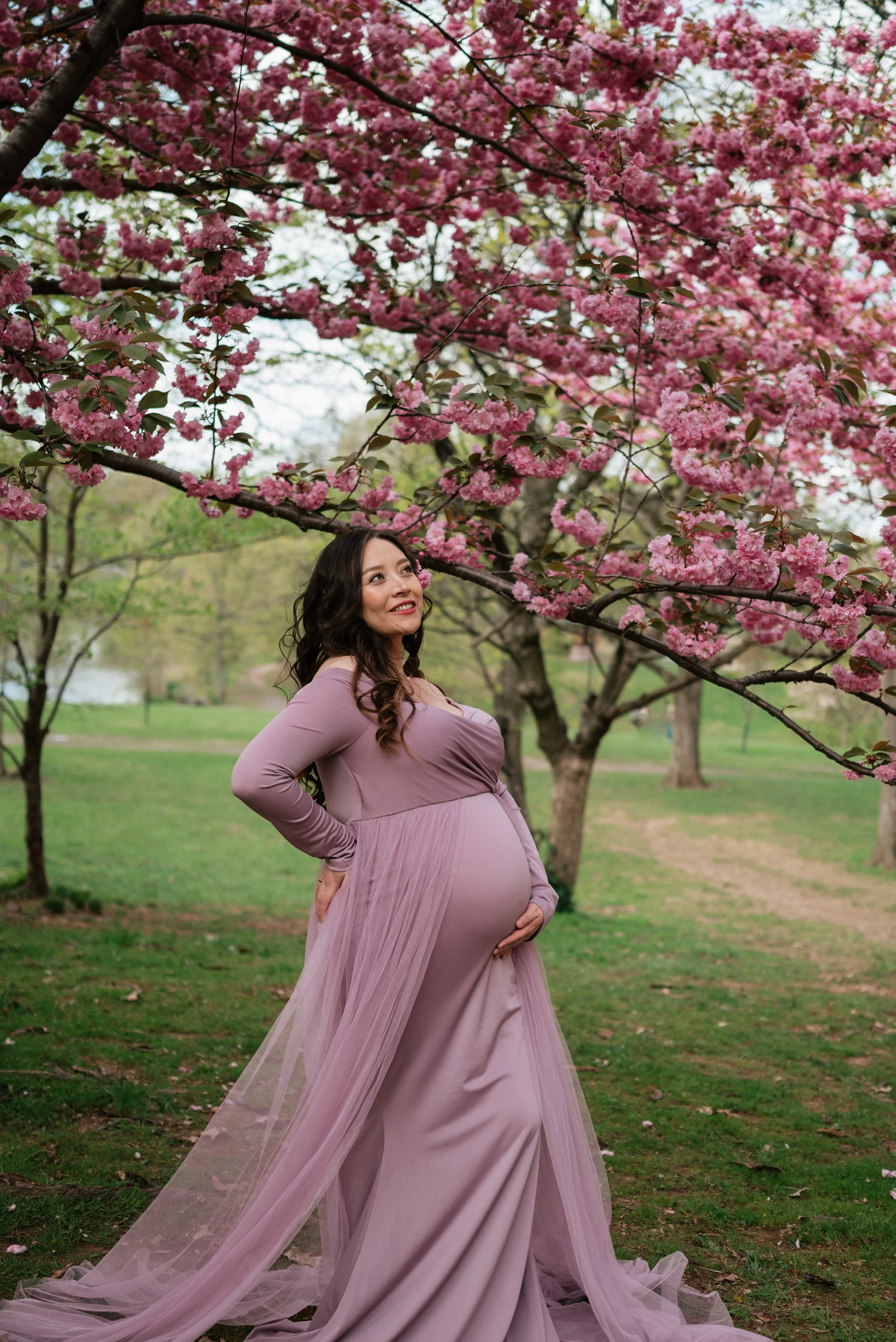 Pregnant woman in a mauve dress standing in a park under pink flowering trees.