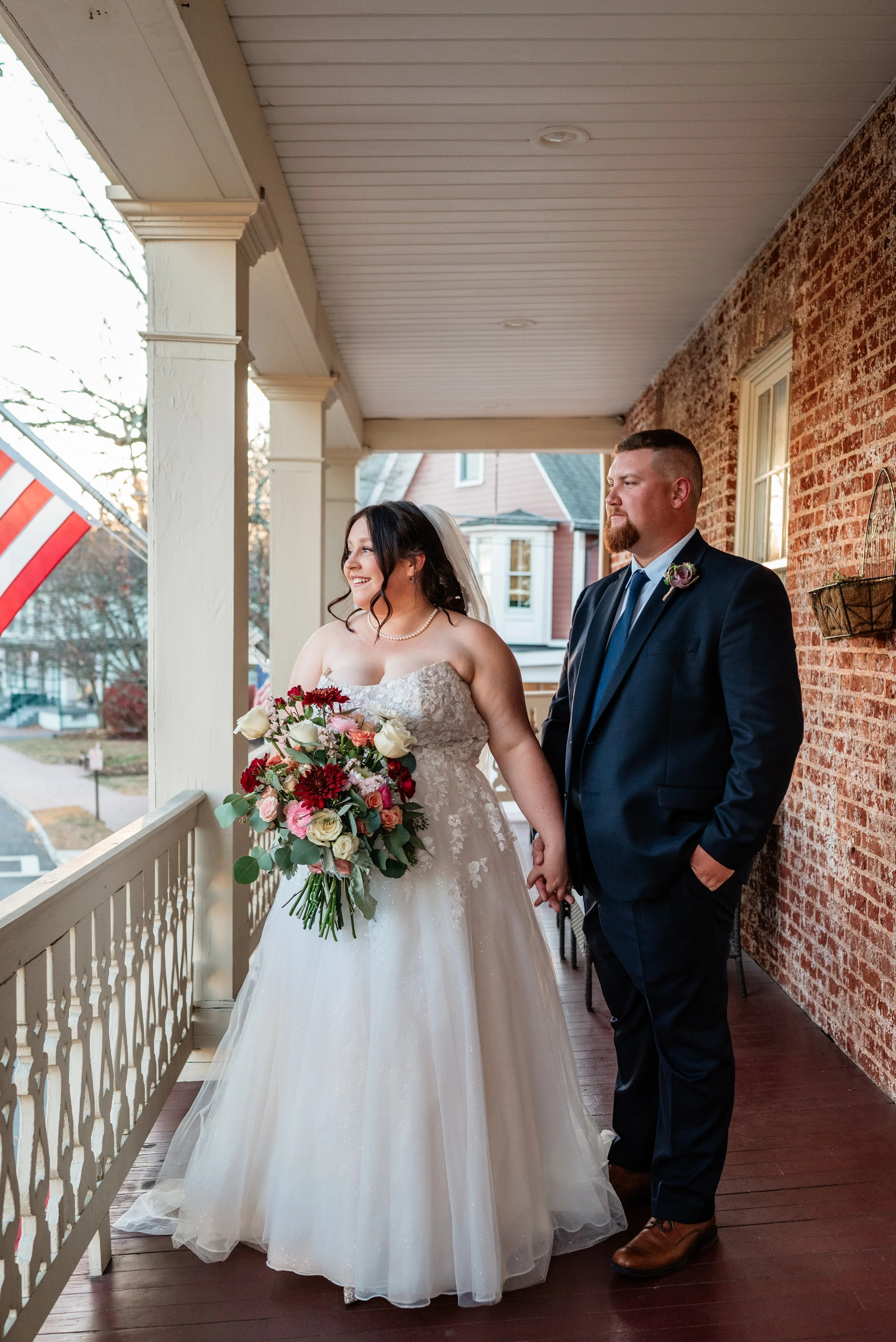A bride and groom stand on a porch, holding hands. The bride wears a strapless white wedding gown and pearl necklace, holding a large bouquet, smiling and looking happy. The groom wears a dark suit with a blue tie and brown shoes, gazing at her. An A