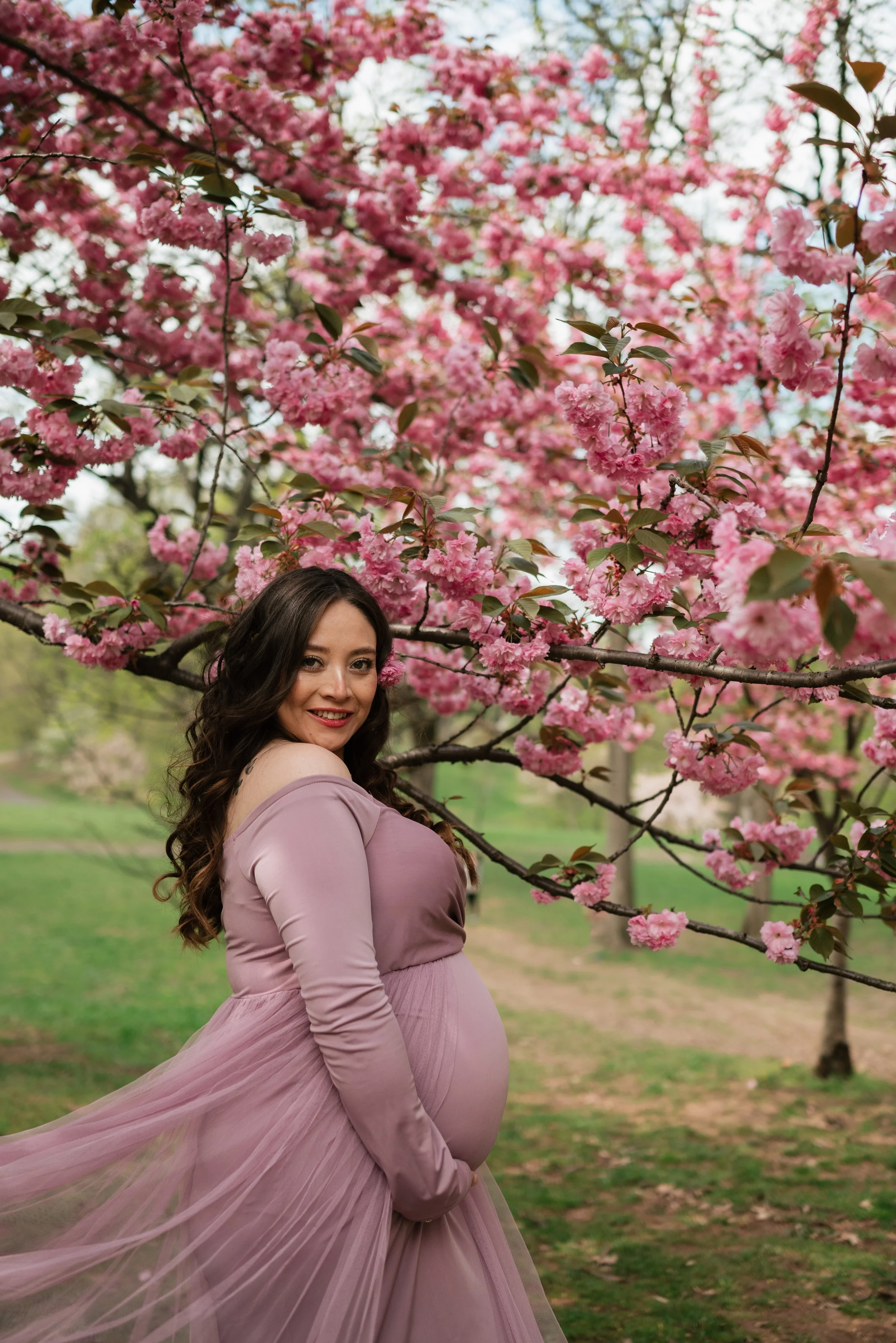 A pregnant woman in a mauve dress standing under pink flowering cherry blossom trees in a park.