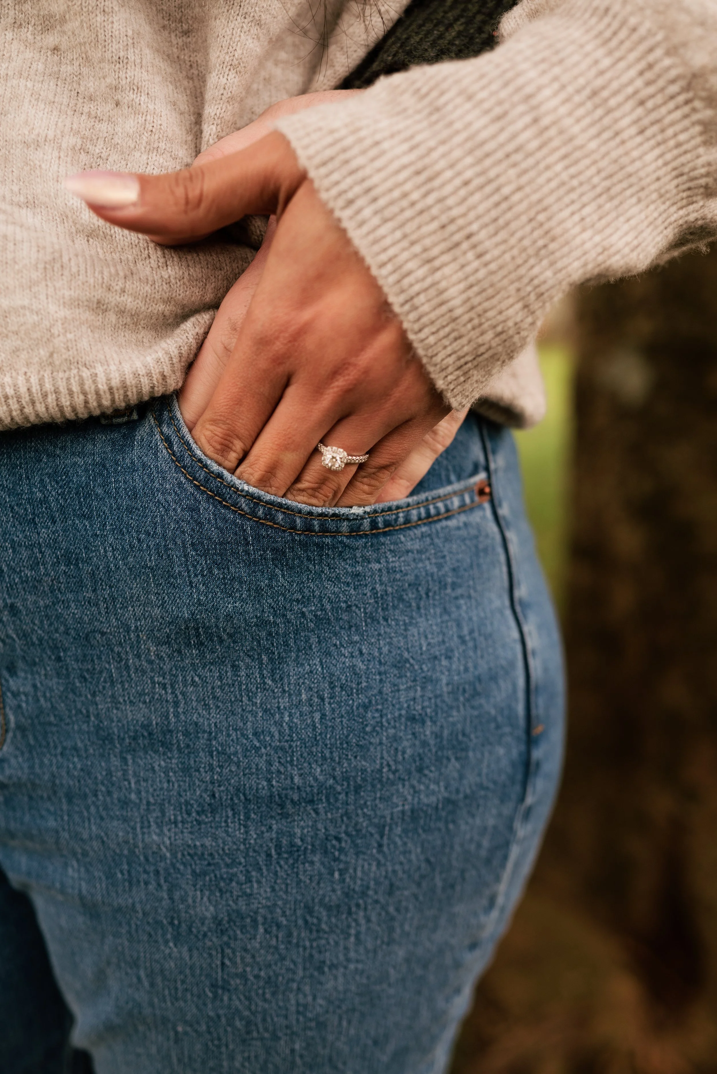 Close-up of a woman's engagement ring, wearing a beige sweater and blue jeans, with her left hand resting in her pocket. The woman is wearing an engagement ring with a large, round diamond on her ring finger.