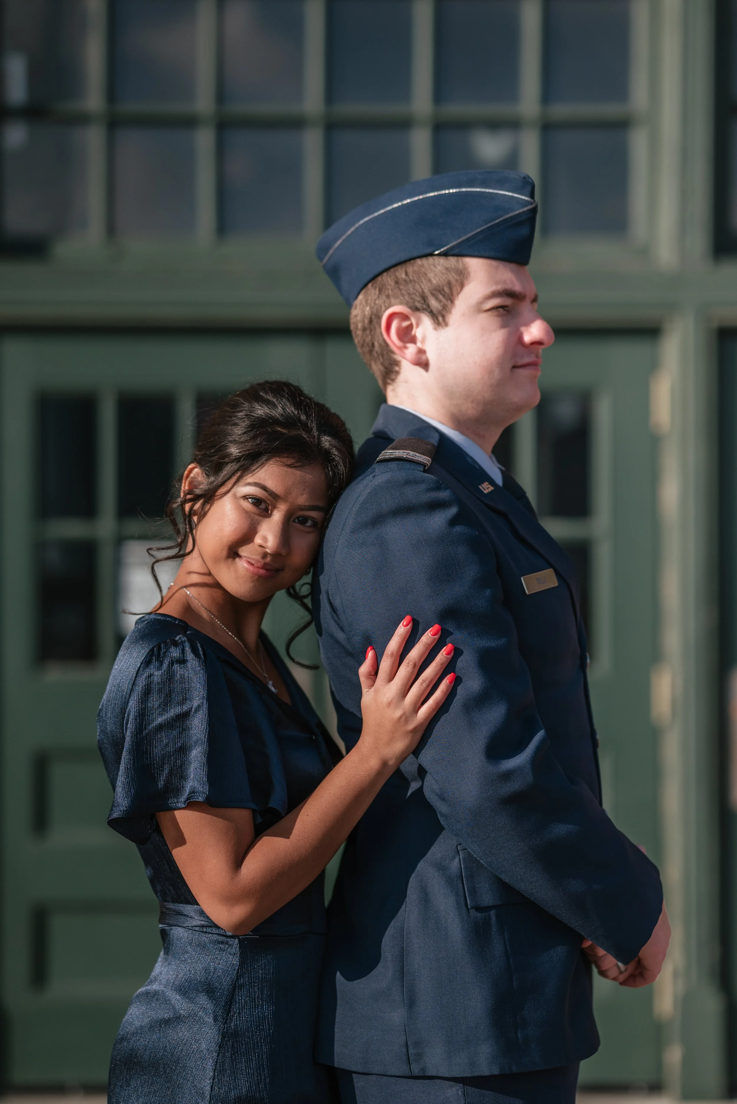 A woman with dark hair and a blue dress leaning on the shoulder of a man in a United States Air Force uniform, standing outside a green building.