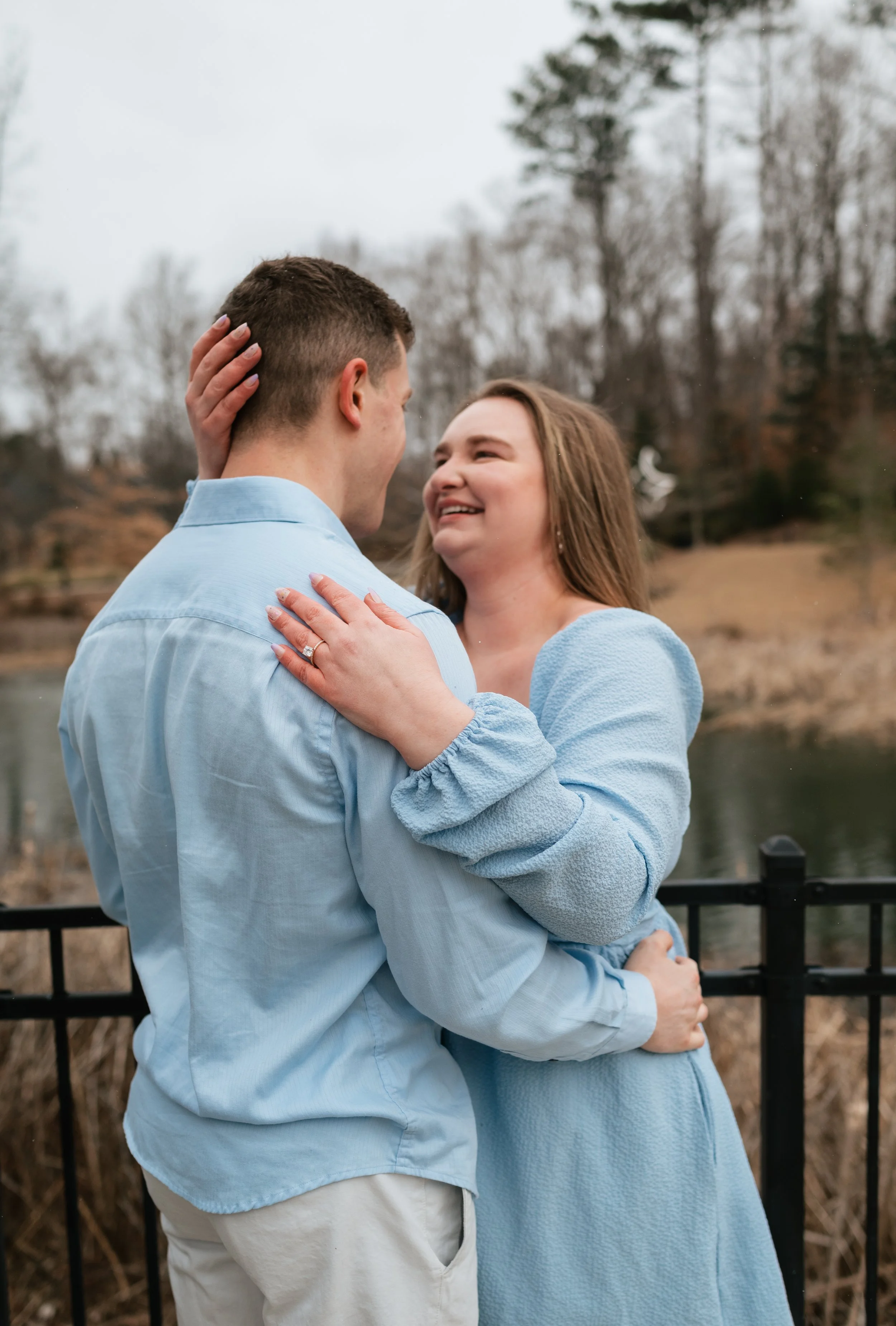 A couple standing outdoors, gazing at each other lovingly near a black metal fence, with barren trees and a body of water in the background, during an overcast day.