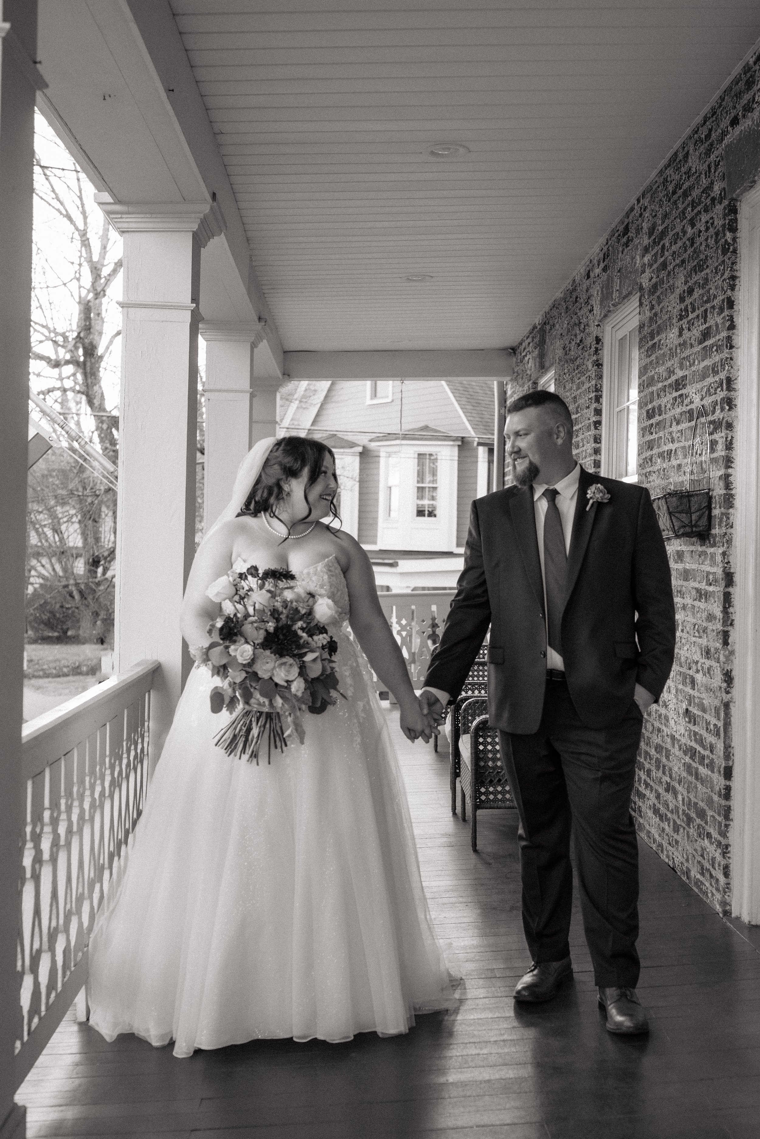 Black and white photo of a bride and groom holding hands on a porch, smiling at each other, with the bride holding a bouquet of flowers.