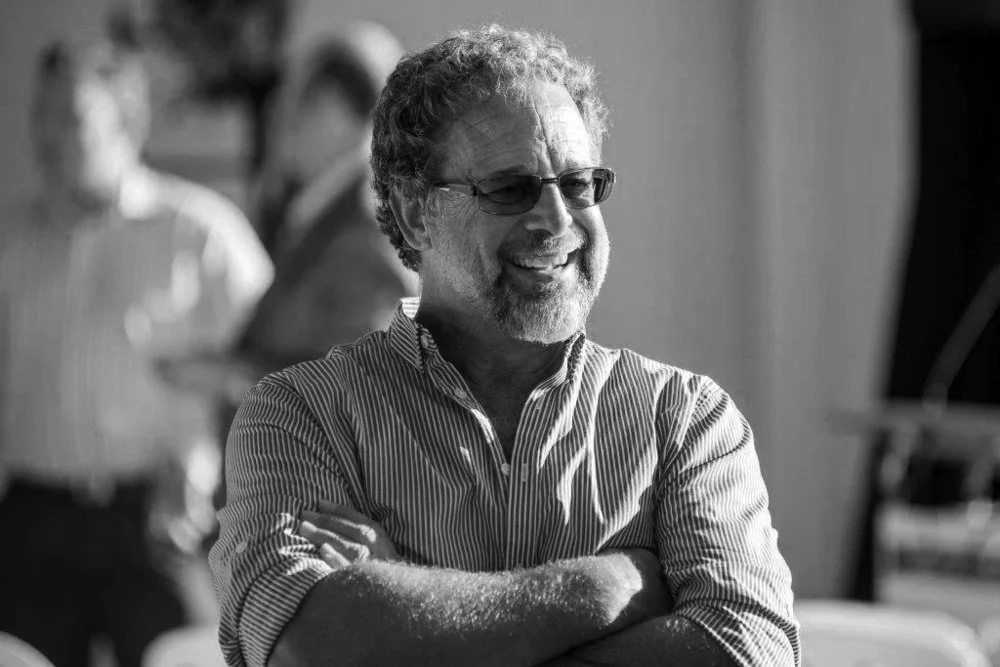 Man wearing glasses with curly hair and cropped beard stands with his arms folded.