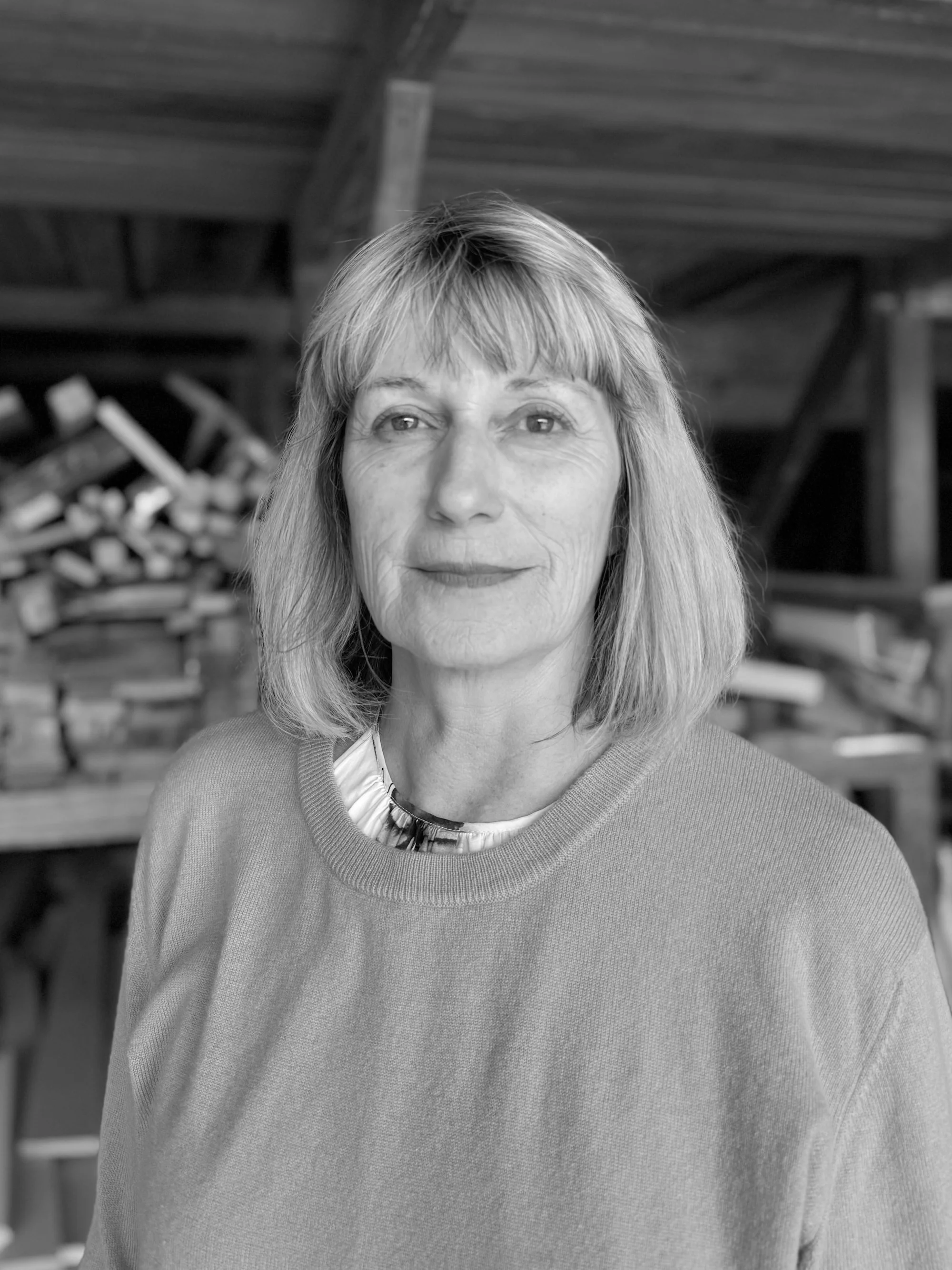 A black-and-white portrait of a woman with shoulder-length blonde hair, wearing a sweater and a necklace, standing in a woodworking workshop with stacks of wood in the background.