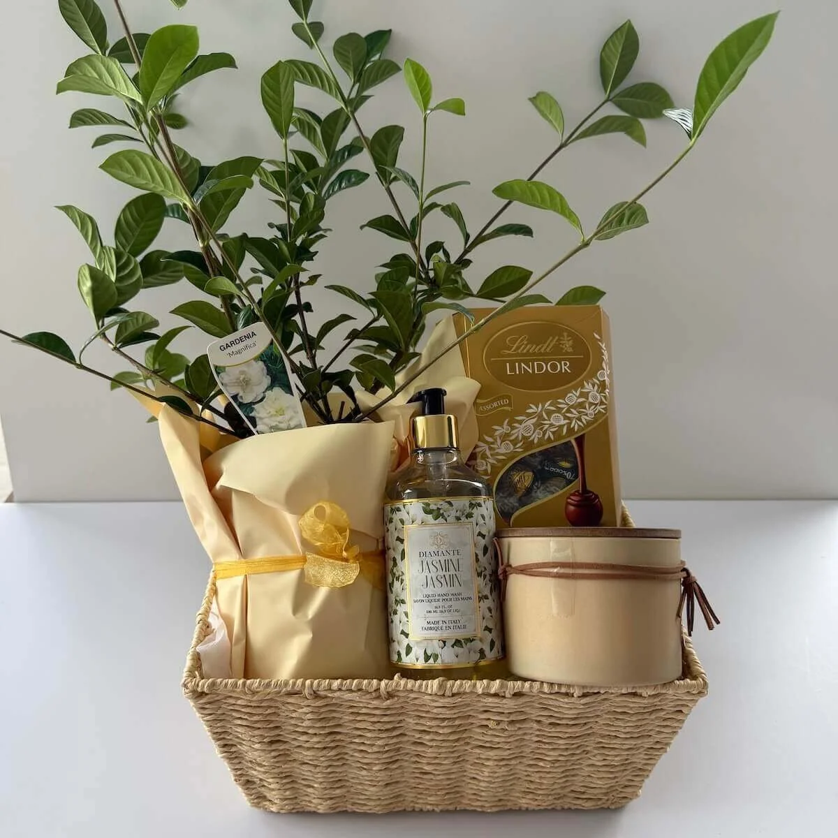 Gardenia Gift Hamper in a woven basket featuring a gardenia plant, scented candle, hand wash, Lindt chocolates, and gift card, beautifully wrapped in cellophane with ribbon for gifting