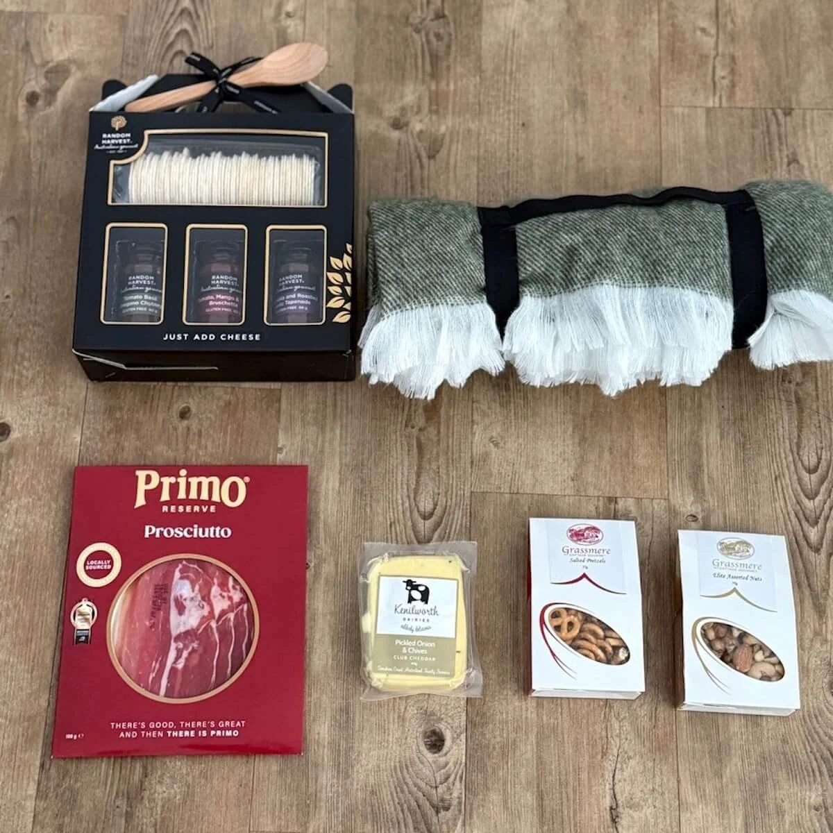 Gourmet picnic items including Ramdom Harvest crackers, chutneys, tapenade, Kenilworth cheddar, prosciutto, Grassmere nuts and pretzels, plates, cutlery, and cups arranged on a flat surface